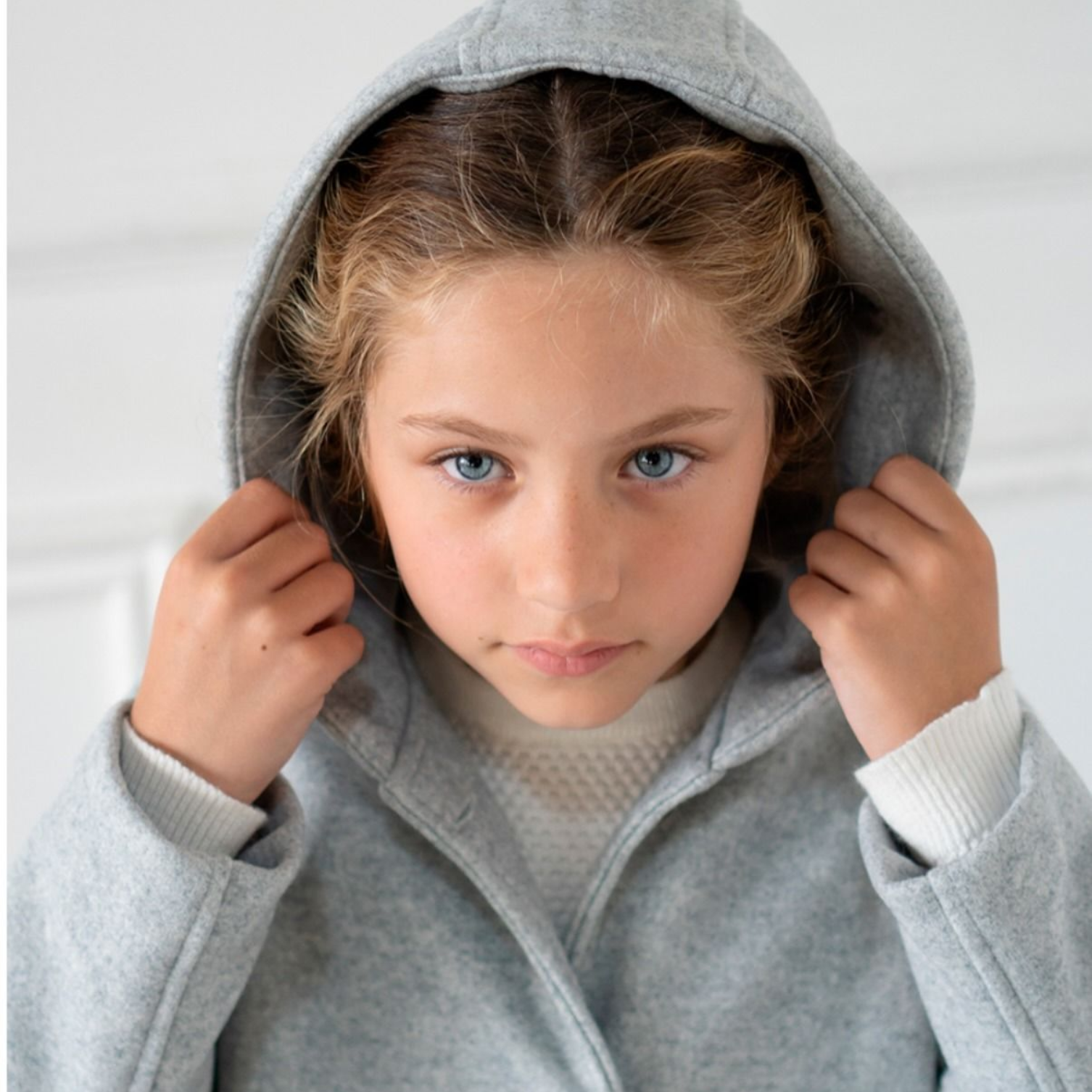 Niña con abrigo gris con capucha, ajustándose la capucha con las manos, expresión seria.