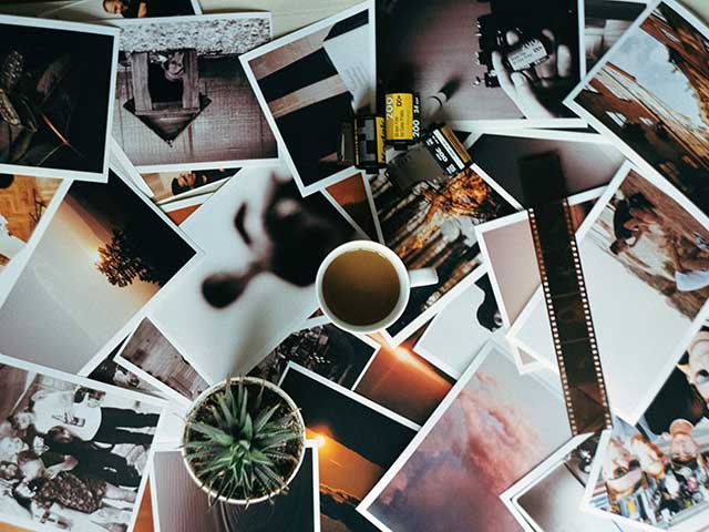 Hundreds of photographs spread out on a table alongside a plant, coffee mug and strip of film.