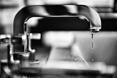 Black-and-white close-up of a kitchen faucet with a single water droplet hanging from the spout