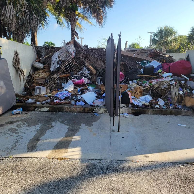 A large pile of debris sits behind a gate in a sunlit outdoor area with palm trees.