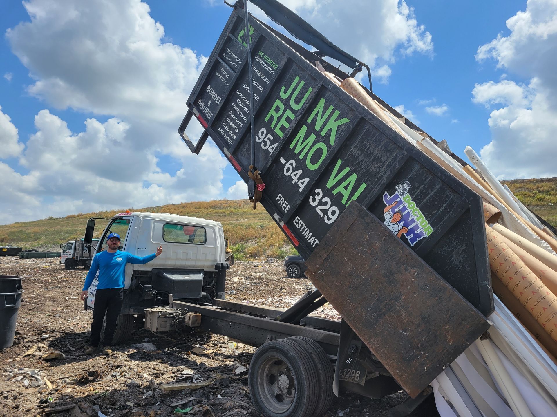 Man in blue shirt points next to a truck dumping junk at a landfill, under a blue sky.