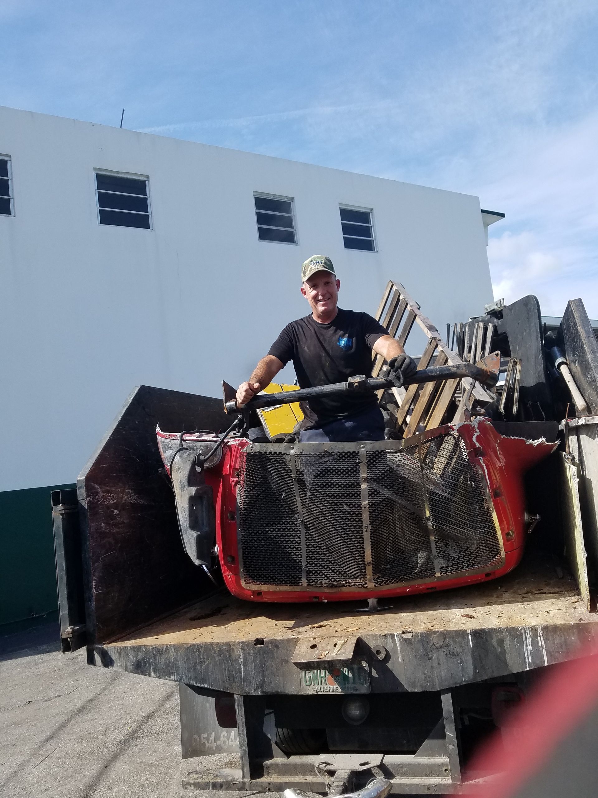 Man in truck bed with scrap metal, smiling in front of a white building on a sunny day.