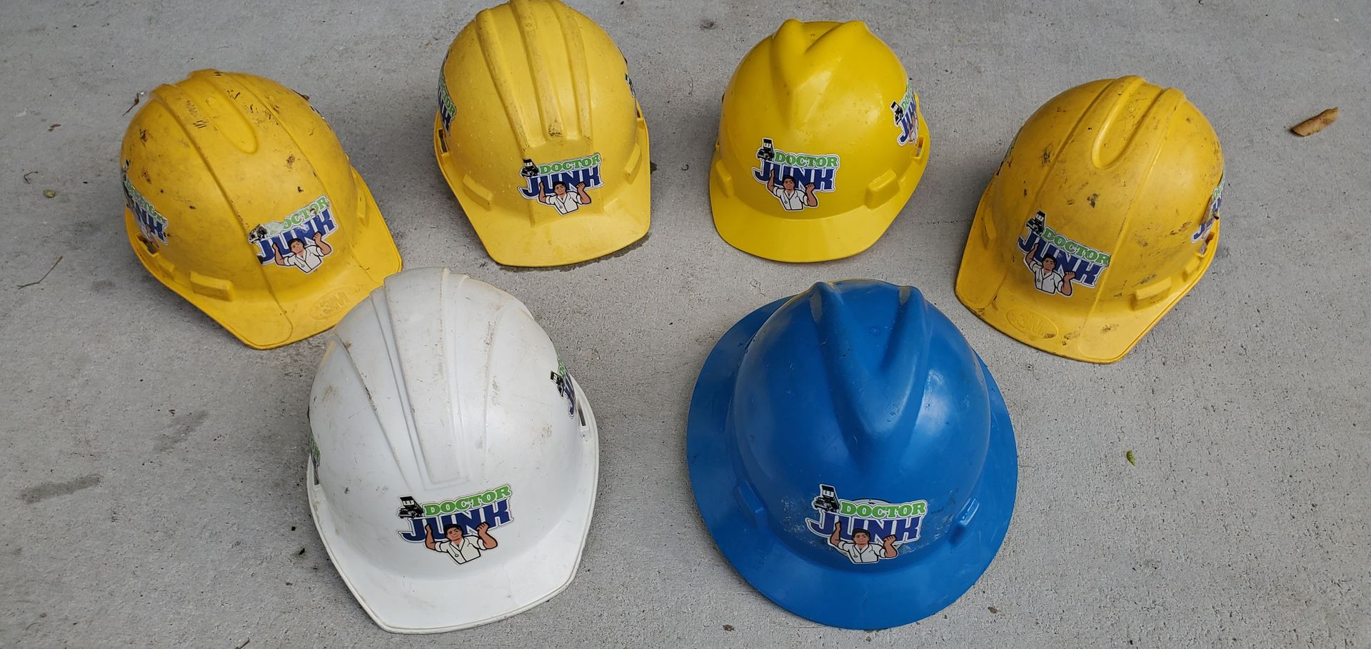 Six hard hats; four yellow, one white, and one blue on a gray surface. Each has a logo on front.