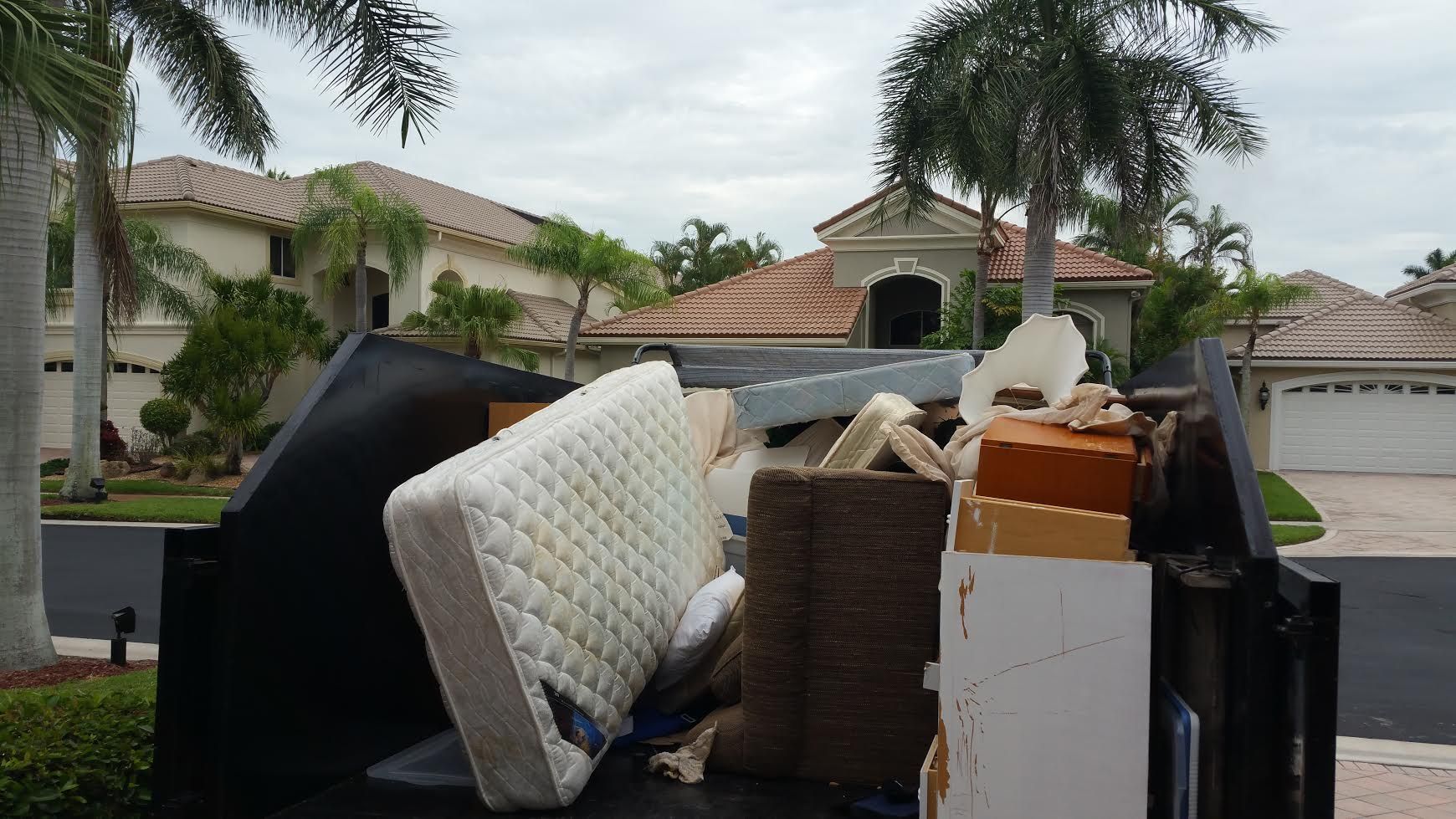 Dumpster overflowing with discarded household items in front of luxury homes.