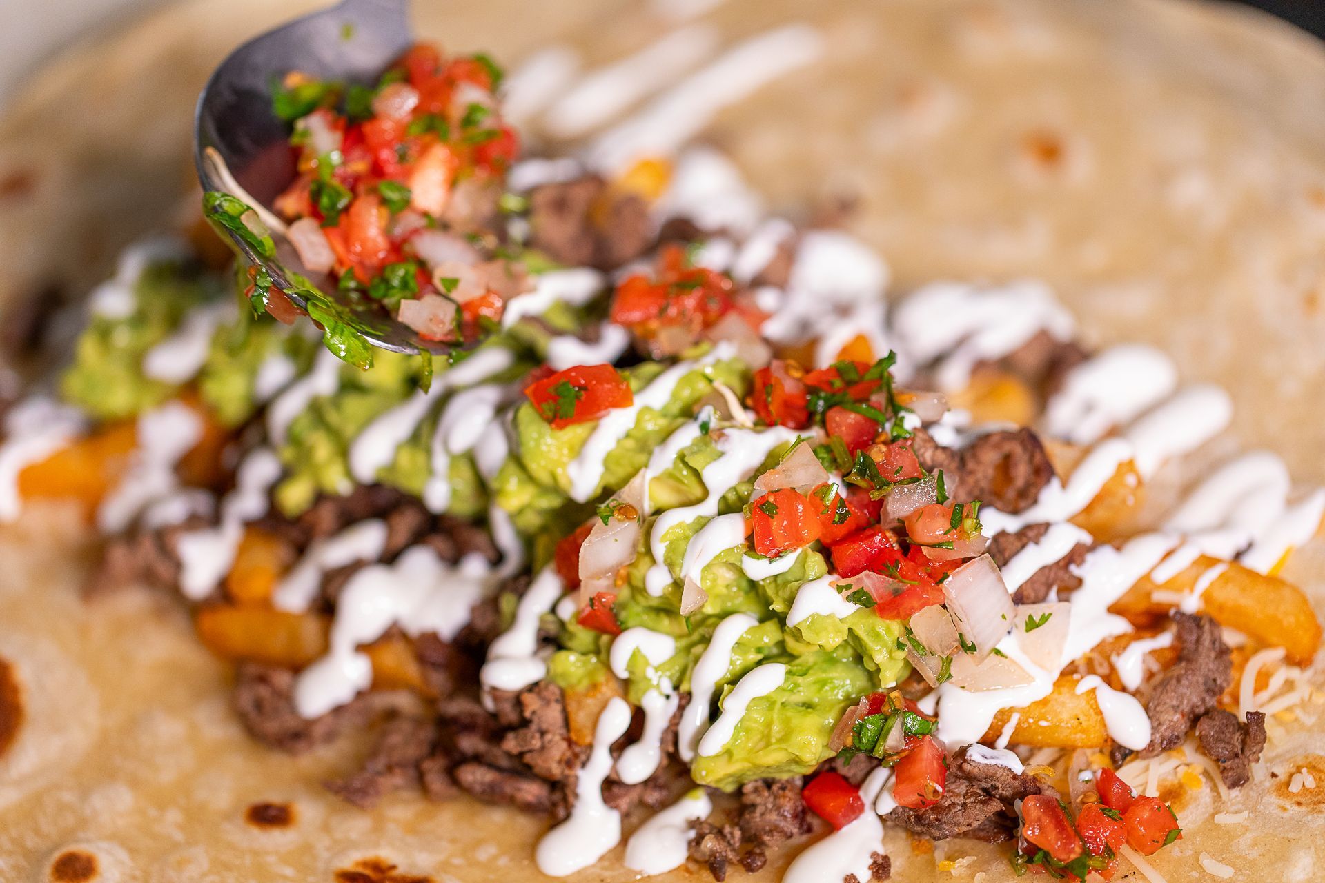 A large burrito being filled with ingredients. Guacamole, meat, fries, and salsa are visible, being added with a spoon.