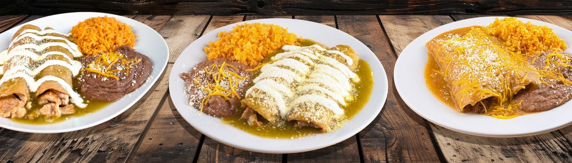Two plates of enchiladas with rice and beans on a wooden table. One plate with white sauce, the other with yellow sauce.