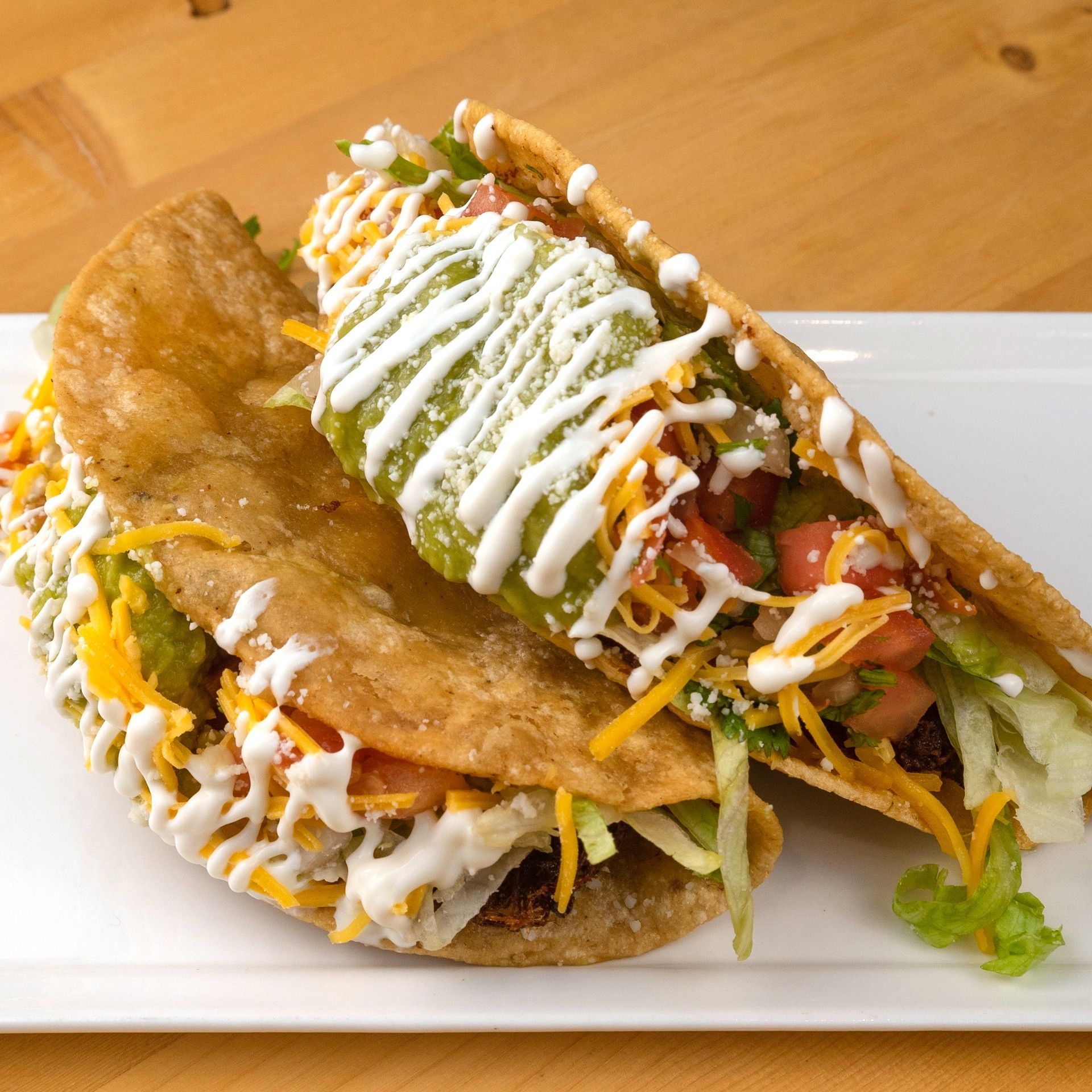 Two crispy tacos on a white plate, filled with lettuce, cheese, tomatoes, guacamole, and cream.