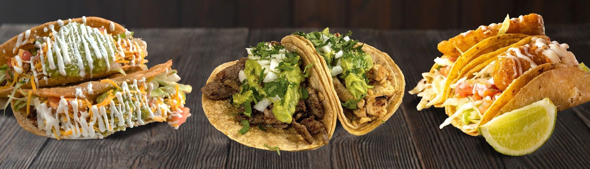 Three tacos with various fillings, arranged on a dark wooden surface, lime slice on the right.