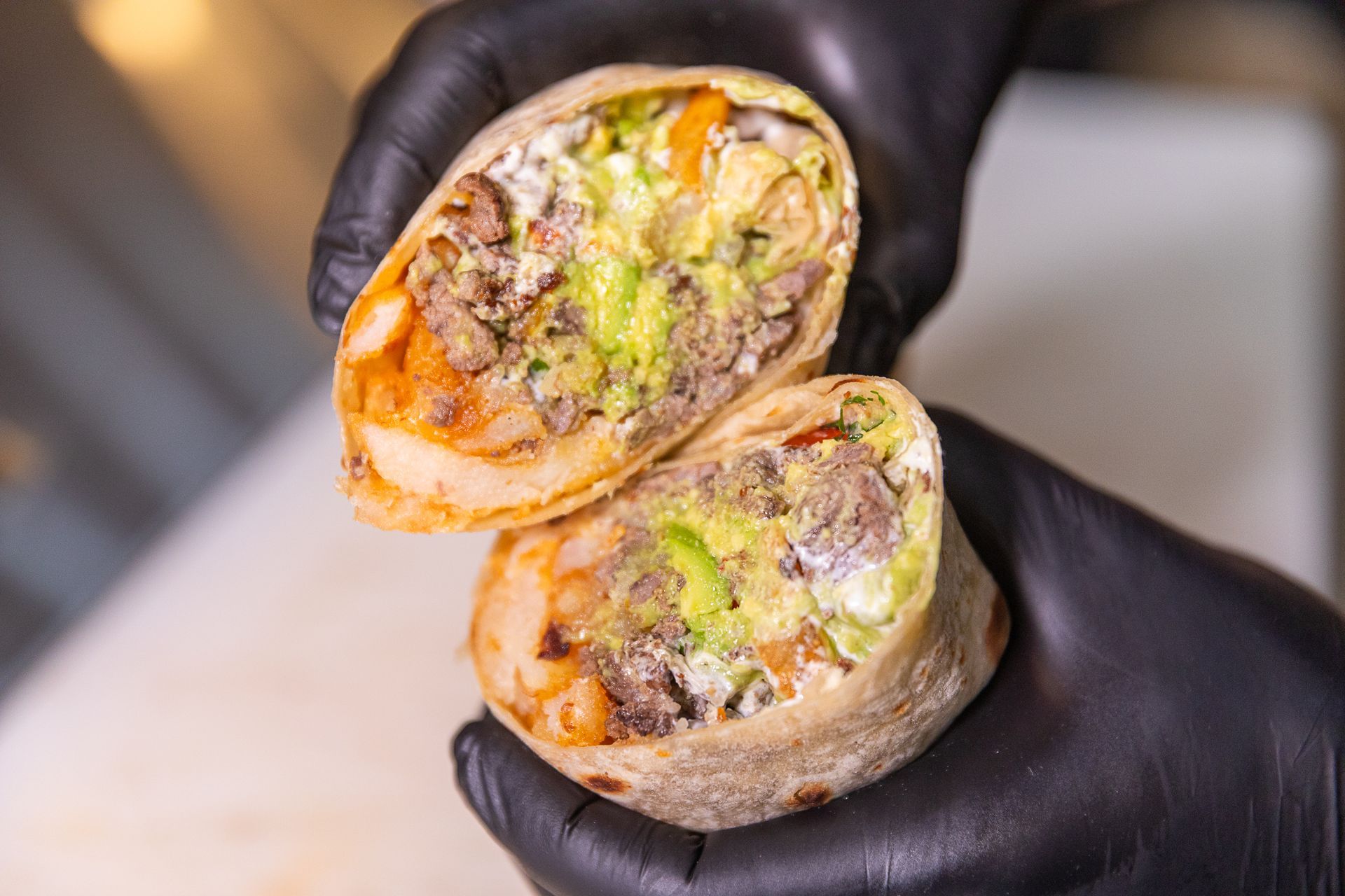 Burrito cut in half, revealing fillings like meat, avocado, and sauce, held by gloved hands.