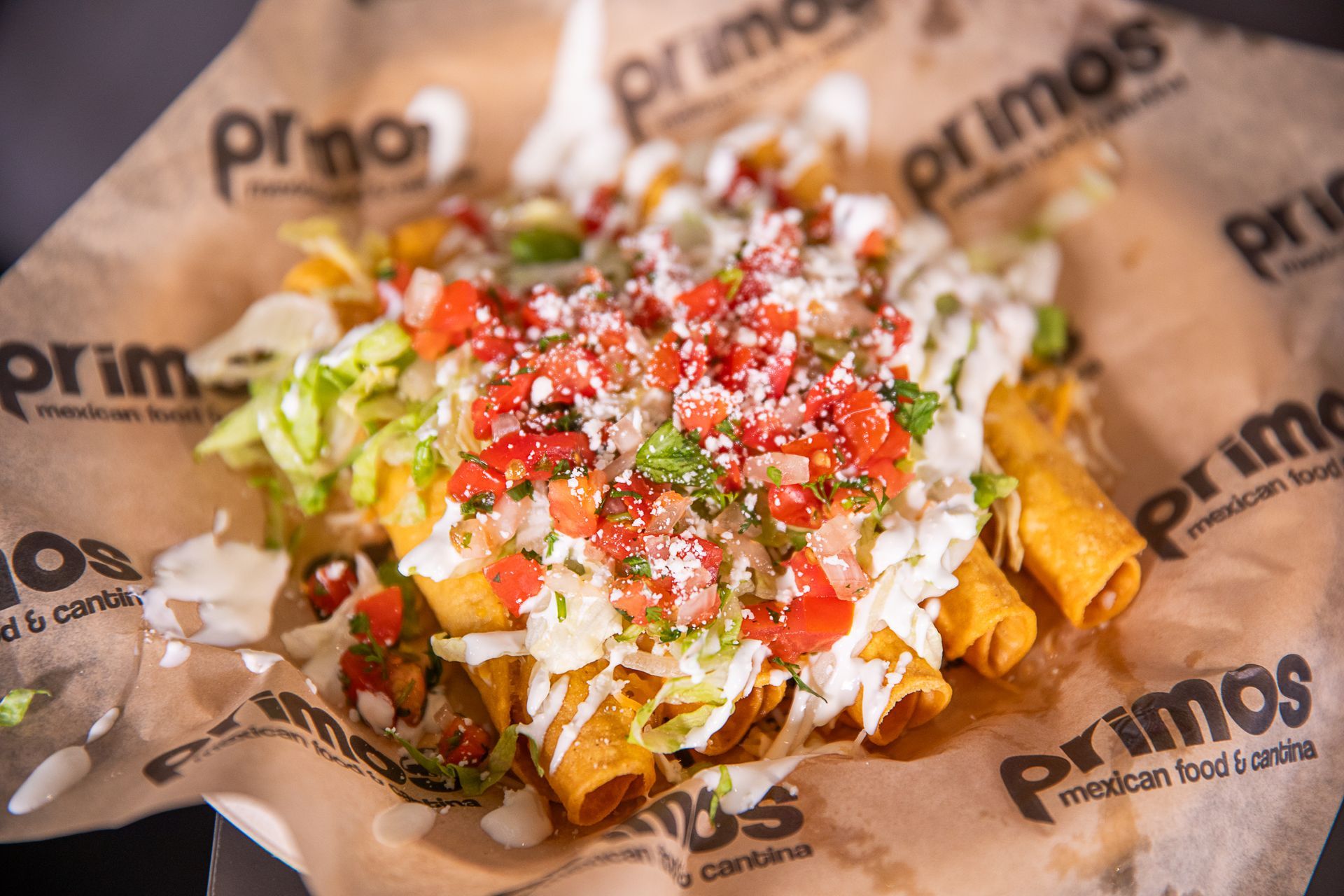 Plate of Primos taquitos, topped with lettuce, salsa, and cream on a printed paper.