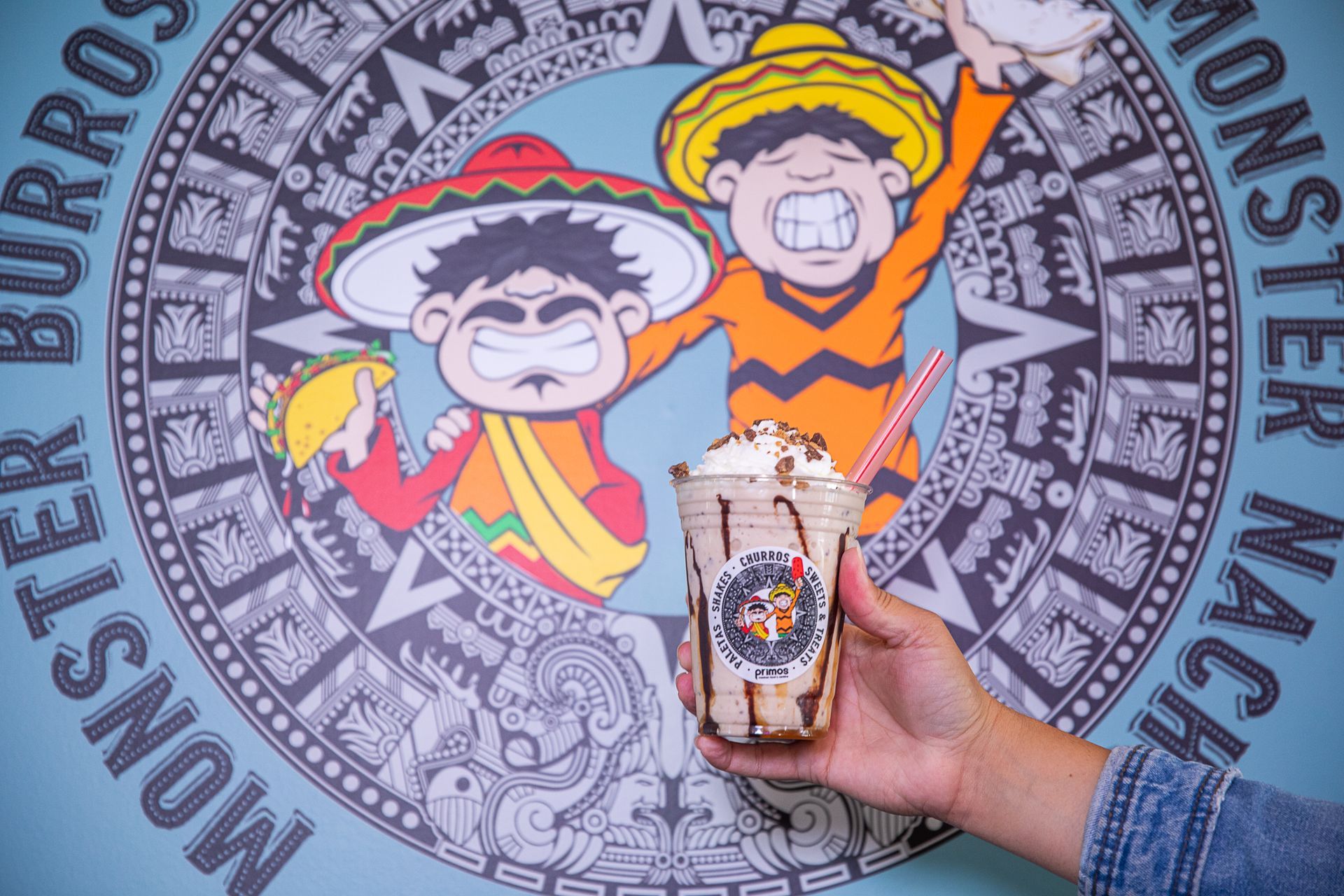 Hand holding milkshake in front of a Monster Burritos logo, colorful cartoon figures with sombreros on a blue wall.