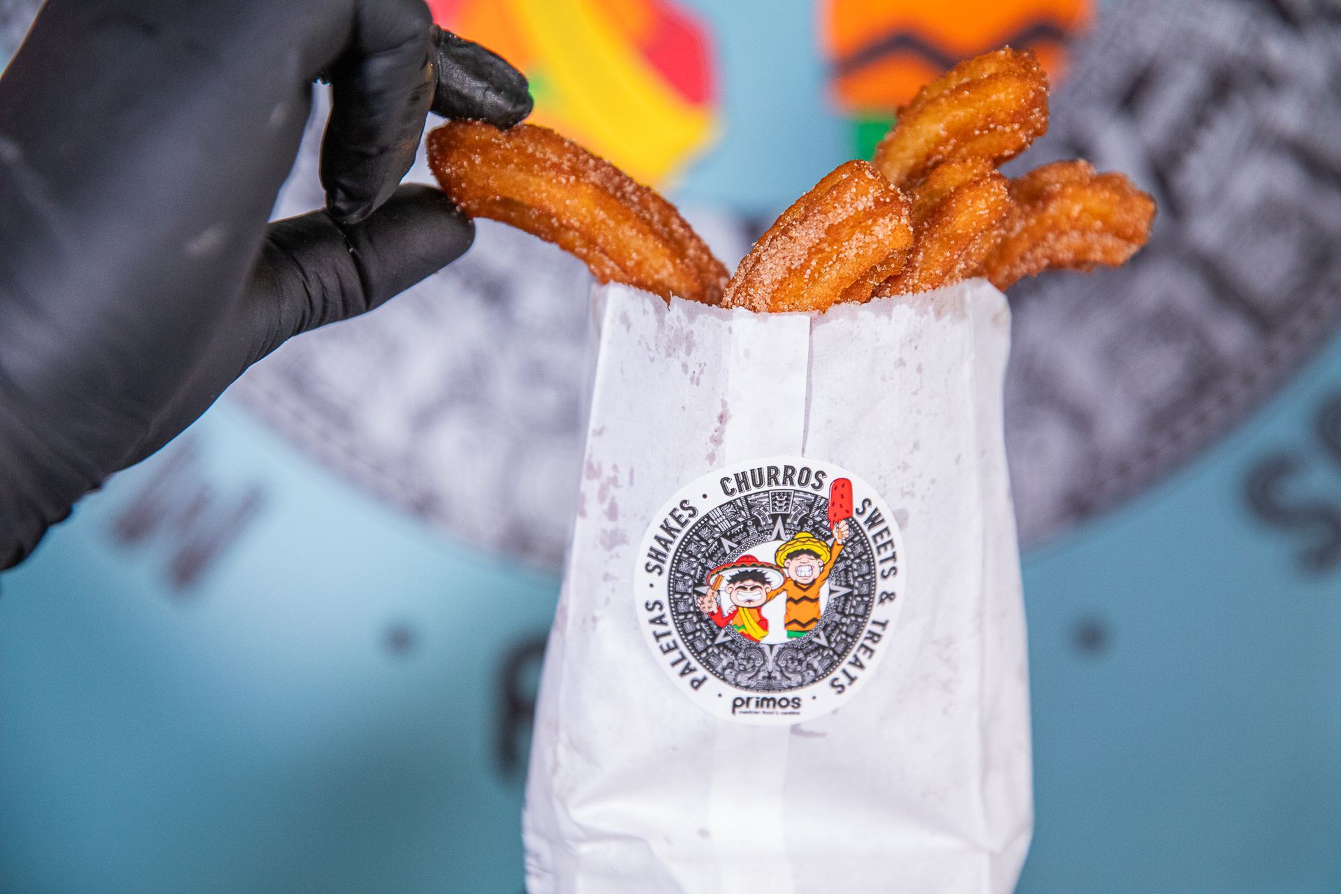 Person in black glove holding churro over a bag of churros. The bag has a logo and is on a blue surface.