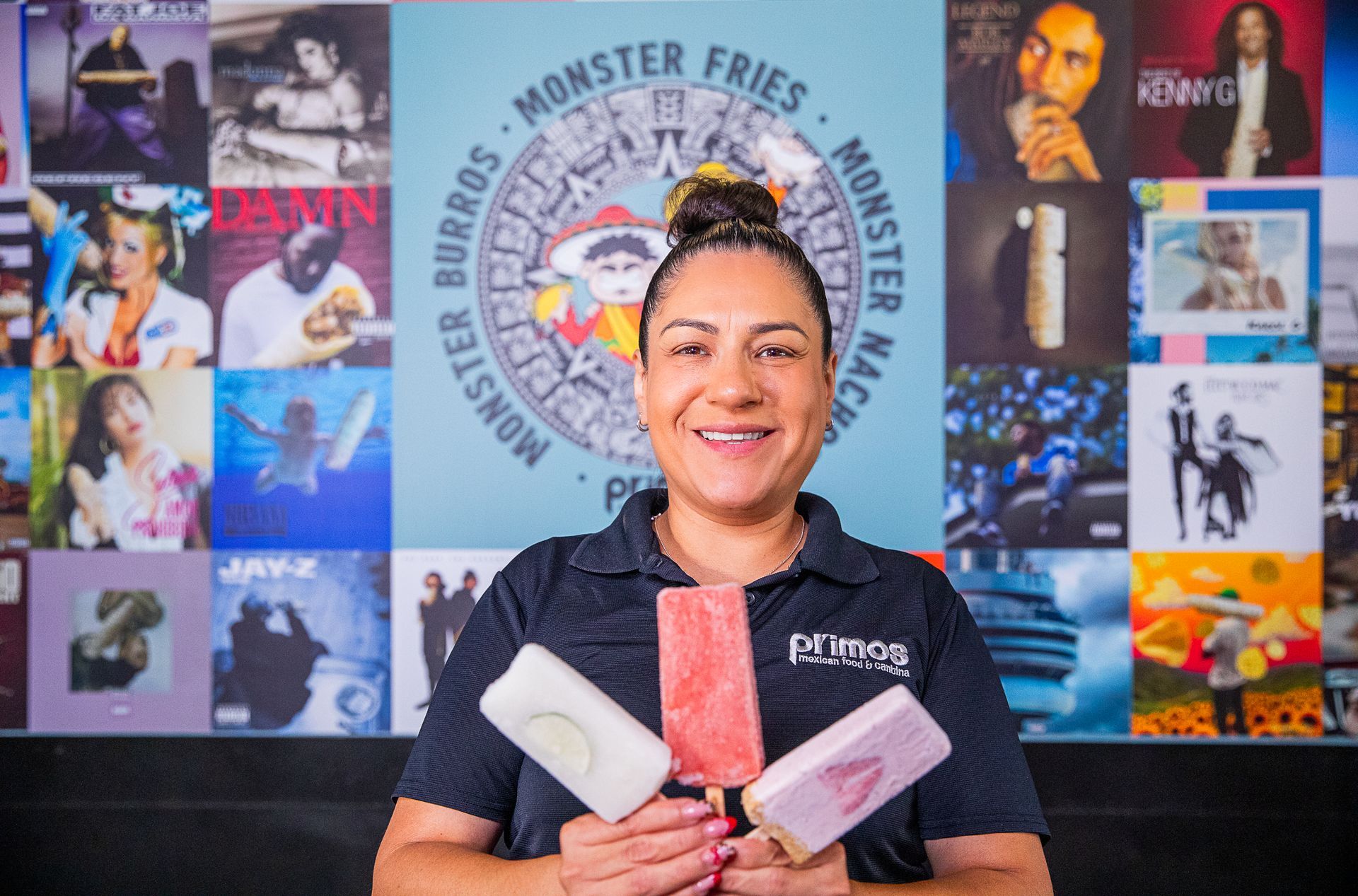 Woman holding three popsicles, smiling, in front of a blue Primo's background with menu text and album covers.