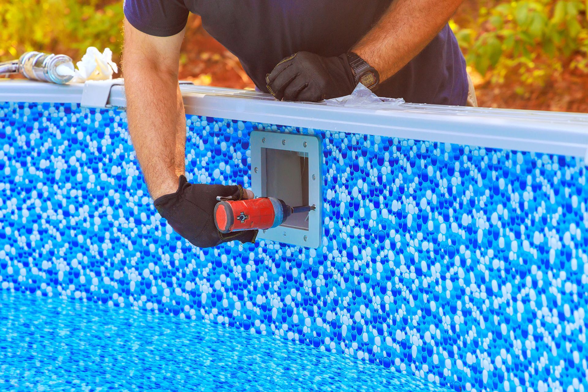 Person in black gloves using a power drill on a pool wall, next to a blue mosaic interior.