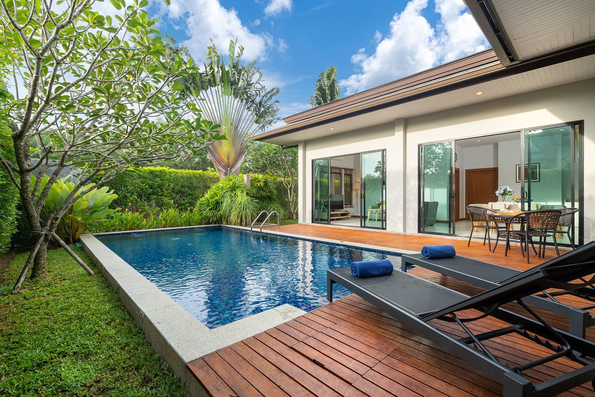 Villa with a pool, sun loungers, and open doors to dining area; lush greenery and blue sky.