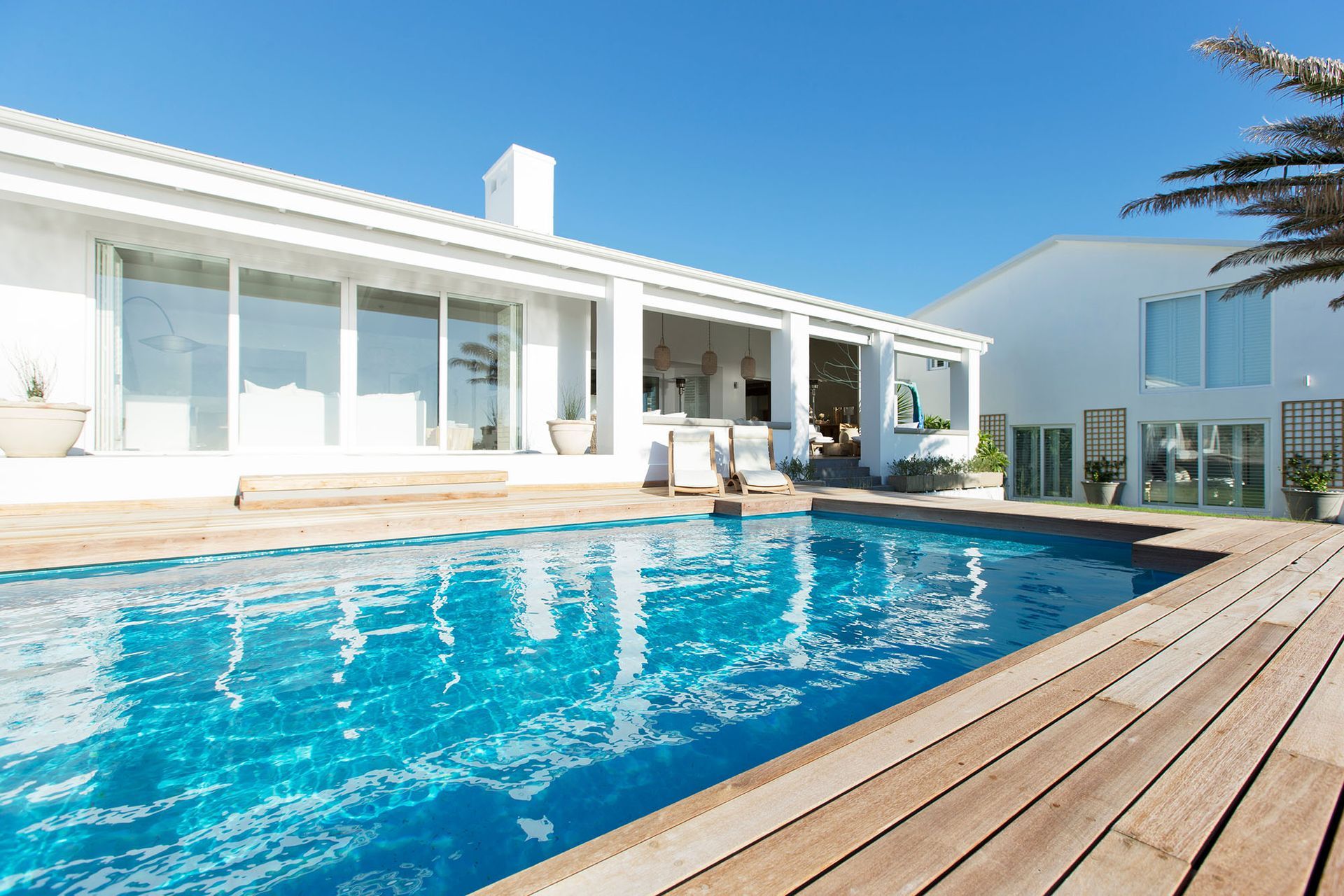 White house with swimming pool, blue water, wooden deck, bright sunny day.