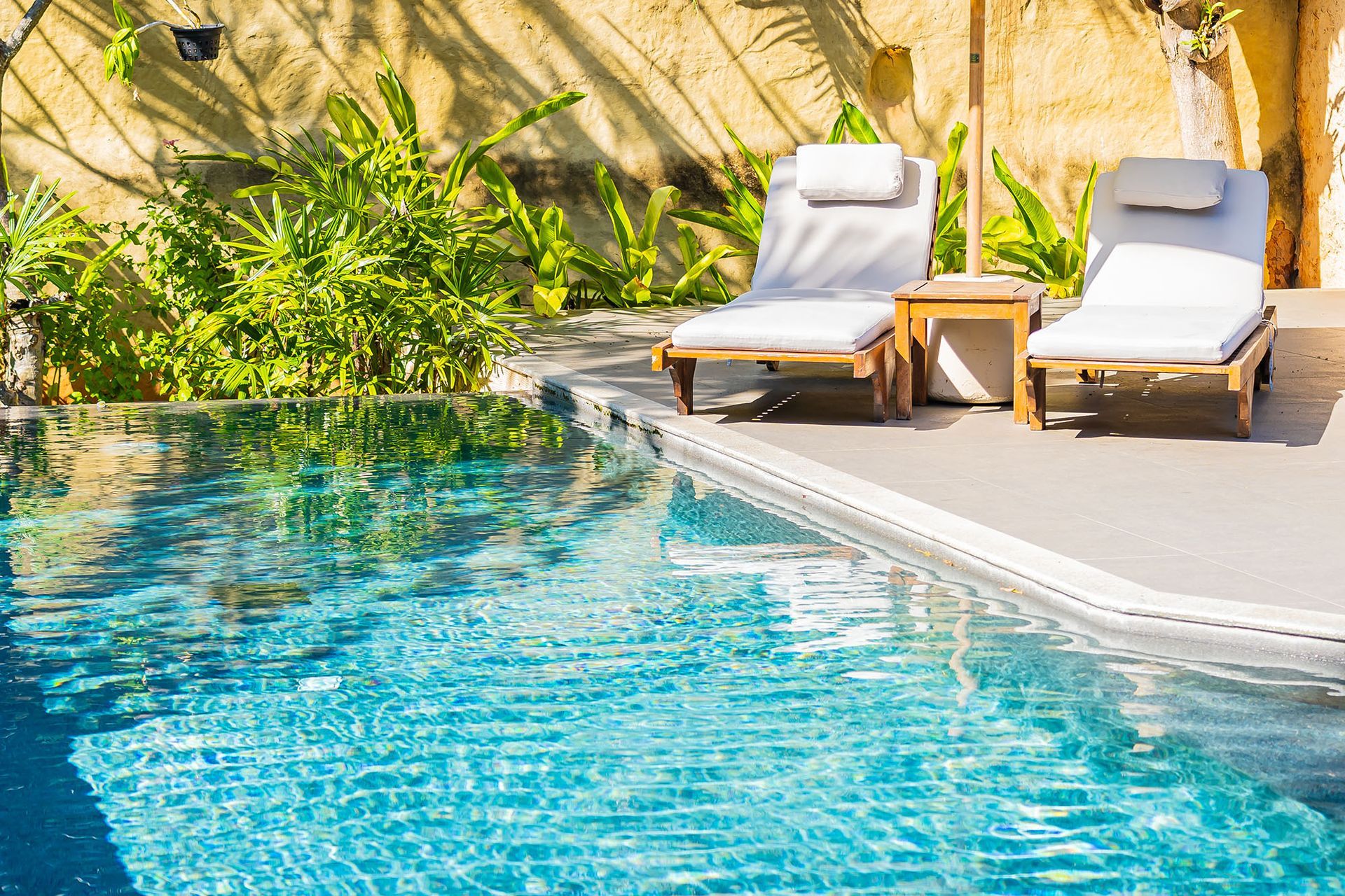 Poolside scene: two white chaise lounges, small table, blue water, greenery, sunlight.