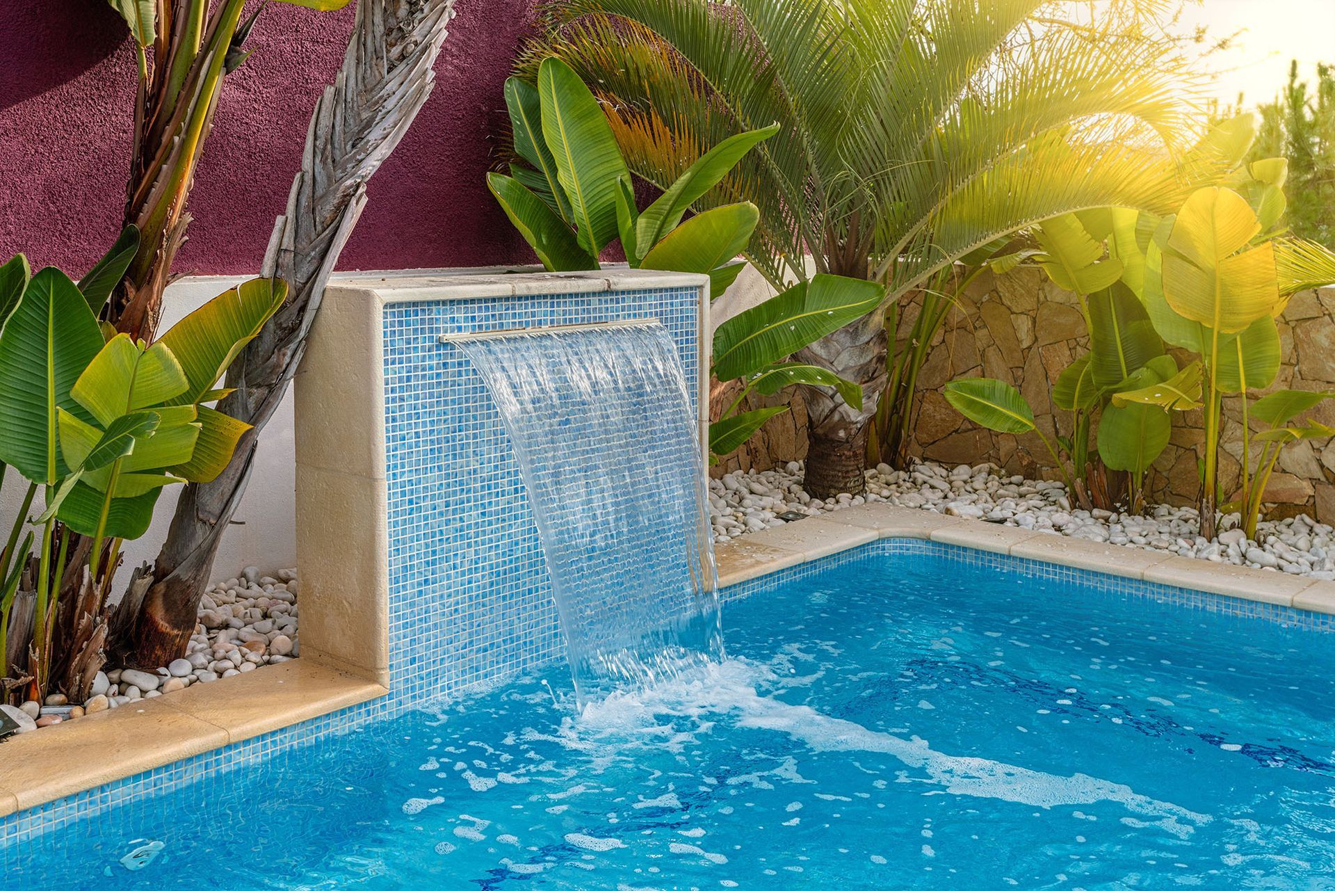Blue tiled pool with waterfall feature, surrounded by lush tropical plants and sunshine.
