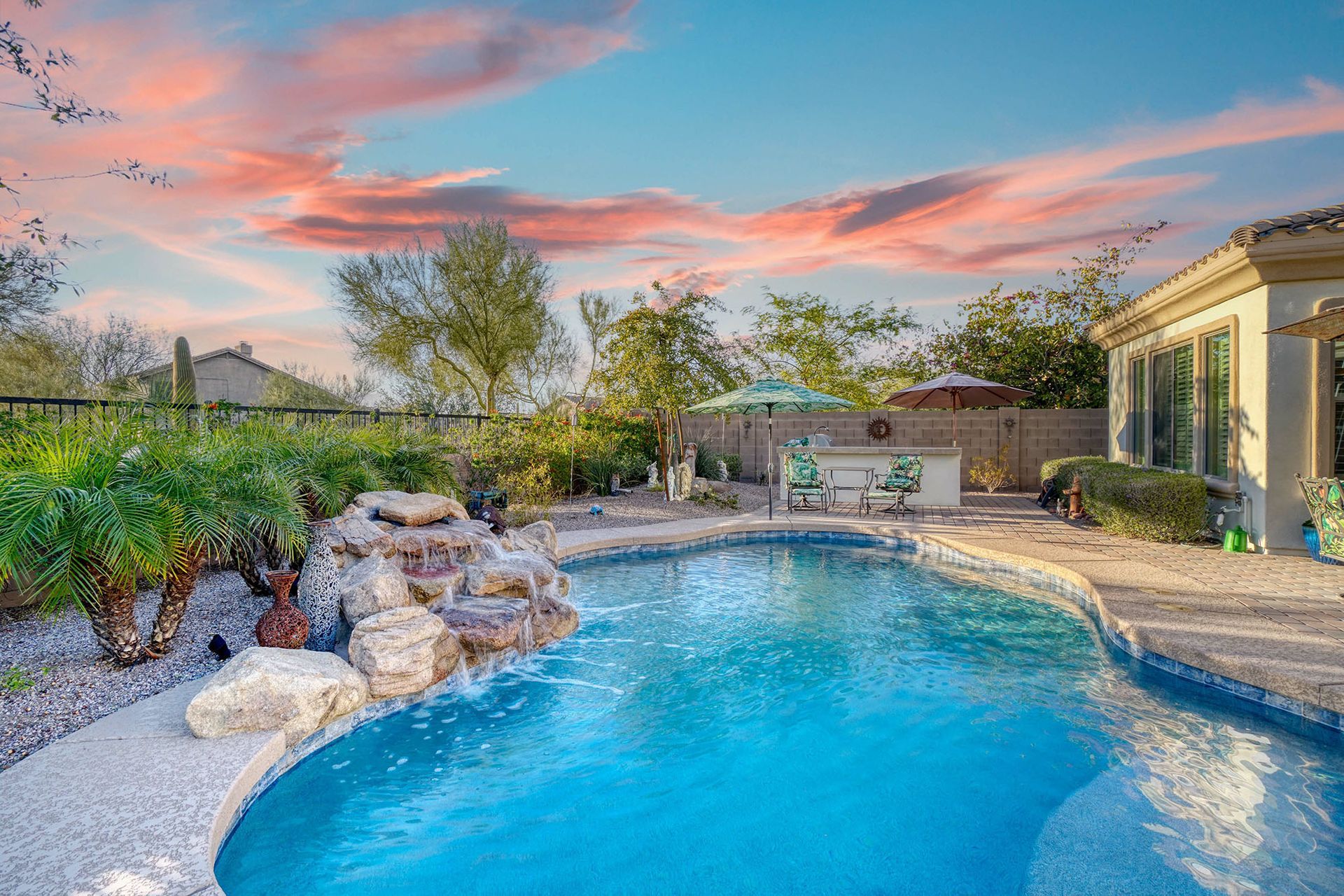 Swimming pool with waterfall feature, surrounded by landscaping, under a colorful sunset sky.