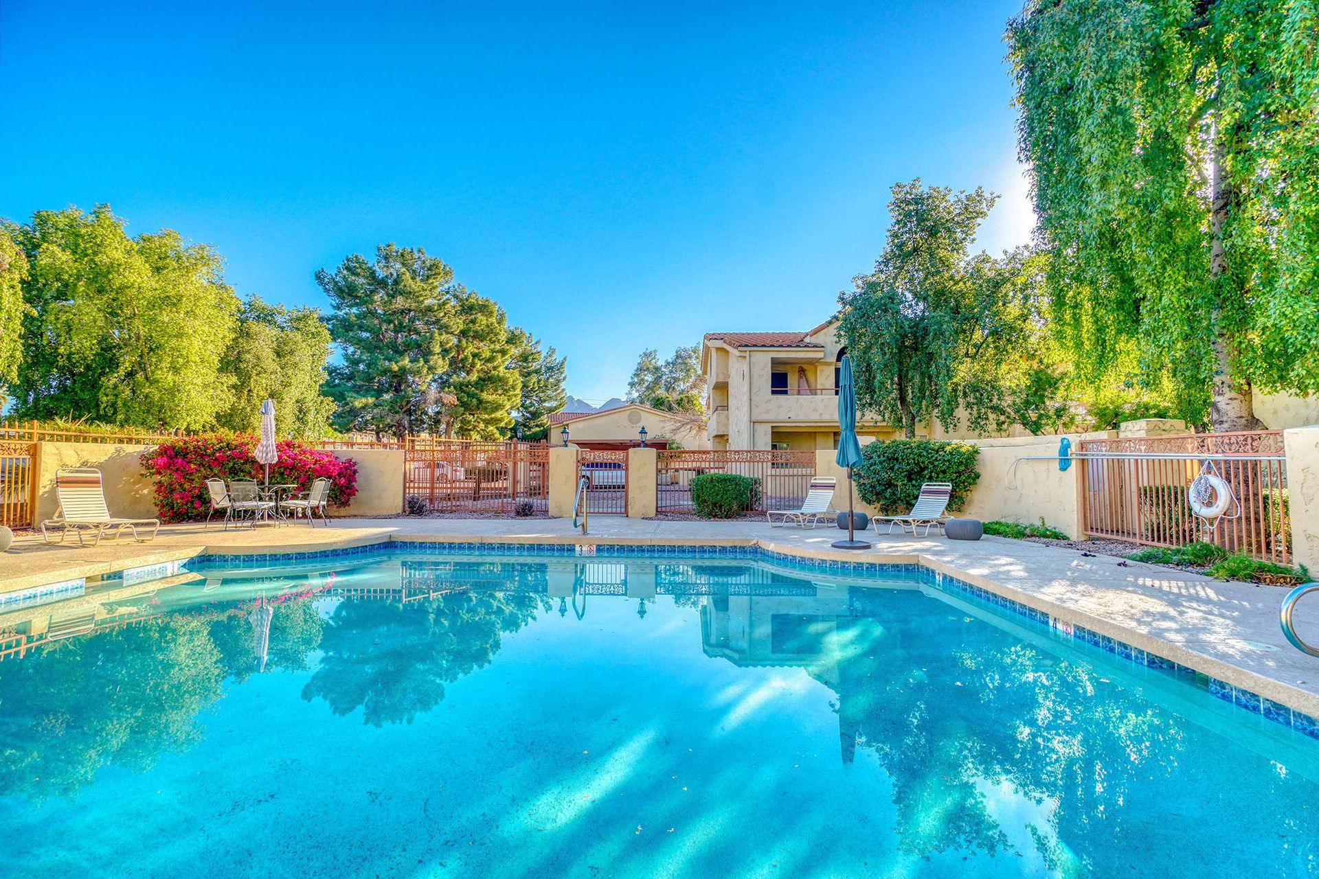 Swimming pool in sunny setting with a two-story building in background. Trees and blue sky visible.