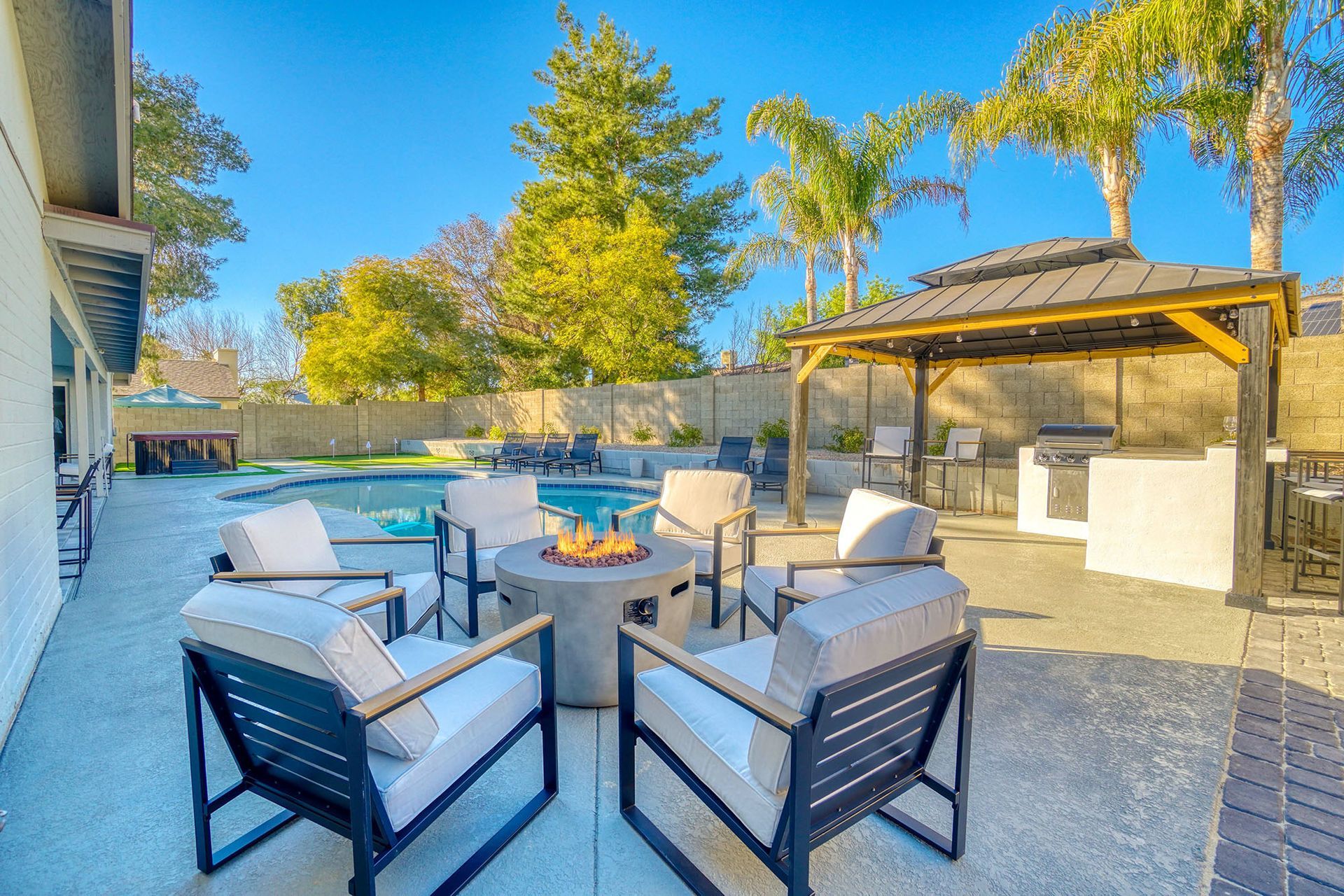 Patio with seating area, fire pit, swimming pool, gazebo, and grill on a sunny day.