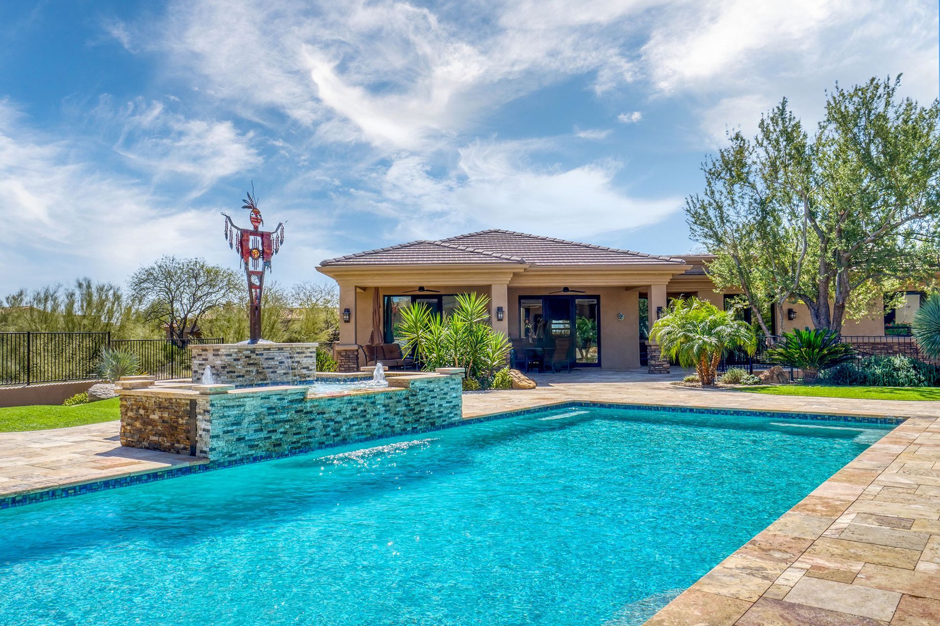 Swimming pool in front of a house, water fountain, sunny day, blue water, tan patio, green lawn, trees, blue sky.
