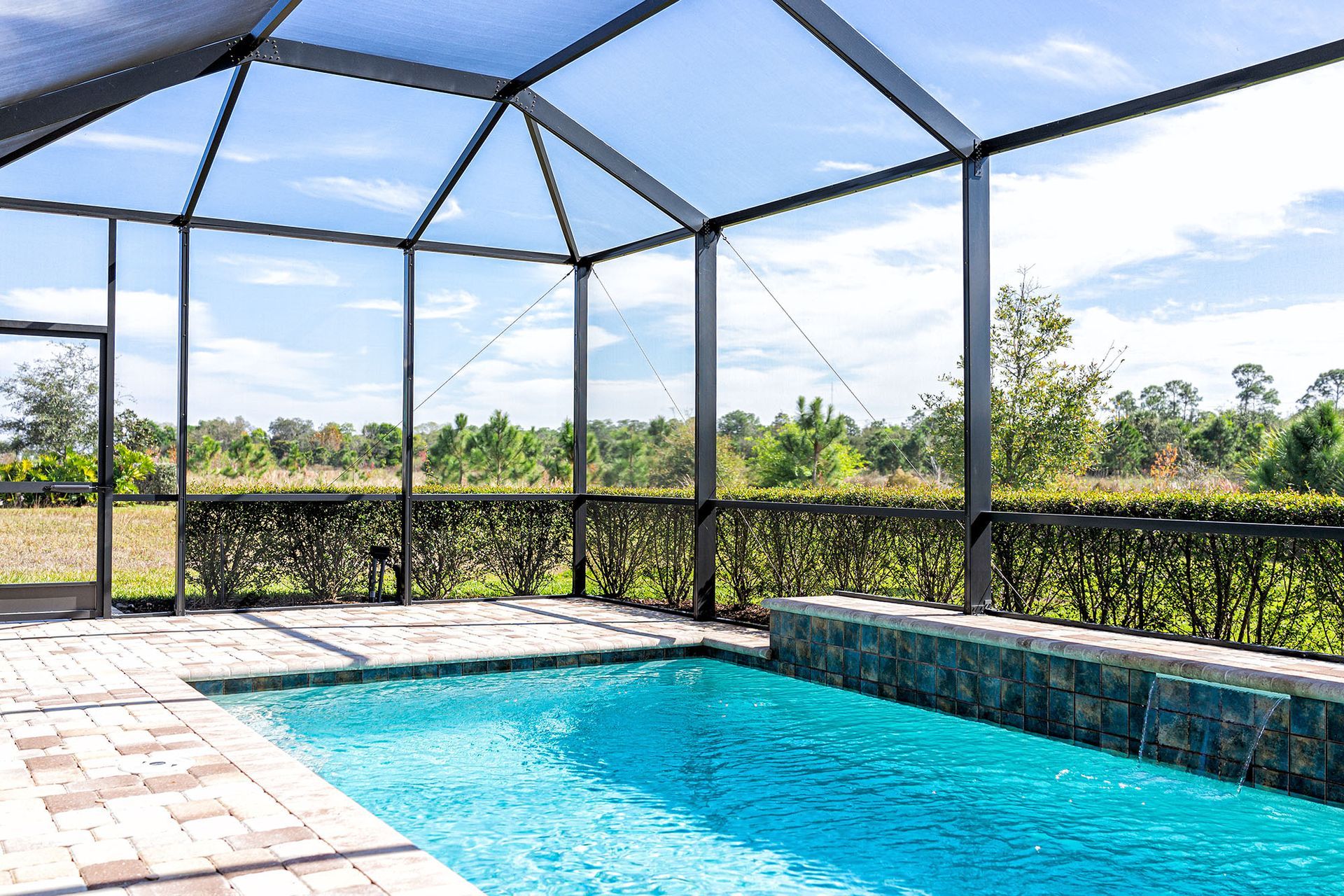 Pool area with screened enclosure, blue water, brick patio, and view of greenery under a bright sky.