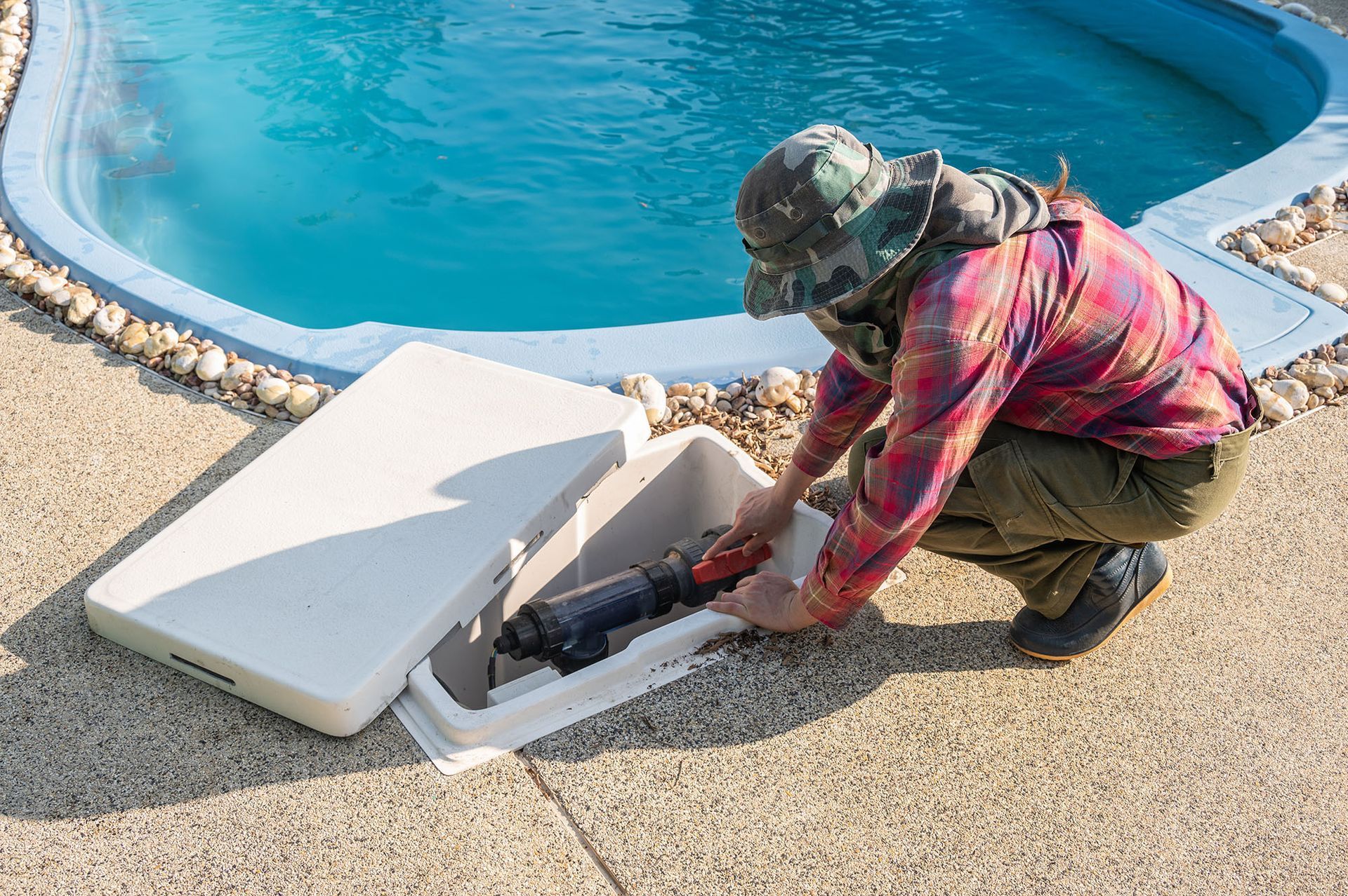 Person opening a white, in-ground pool equipment box, near a pool, retrieving a tool.