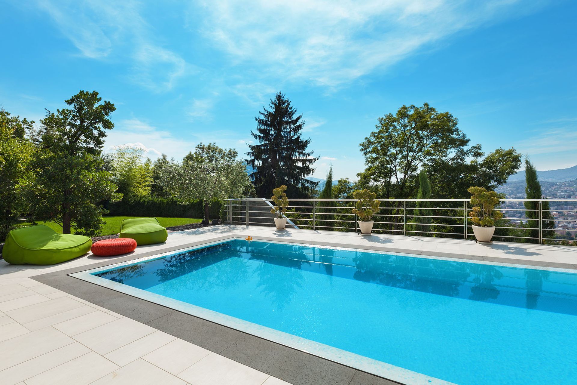 Swimming pool surrounded by a stone patio and lush greenery under a bright blue sky.