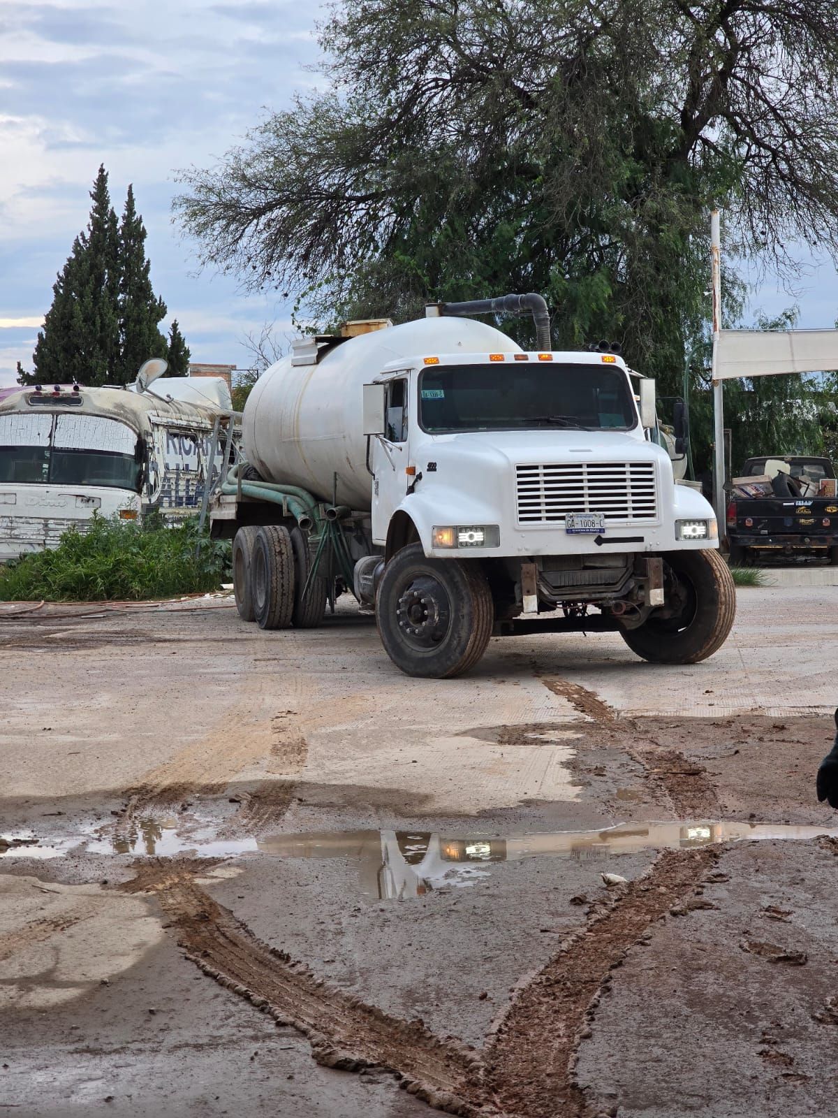 Camión cisterna de agua blanca sobre una superficie fangosa, estacionado cerca de edificios y árboles bajo un cielo nublado.