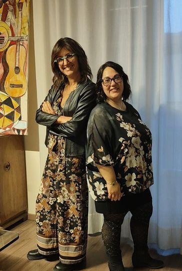 Due donne in piedi, schiena contro schiena, sorridenti. Una in tuta floreale e giacca di pelle, l'altra in camicetta floreale e leggings.