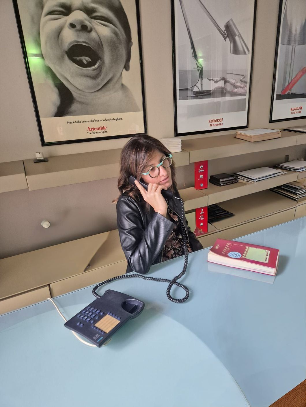 Donna al telefono seduta alla scrivania, con lo sguardo rivolto verso l'alto. Dietro di lei ci sono scaffali con poster. La scrivania è azzurra.