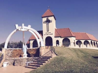 Spanish-style church with red tile roof, tower cross, and white arch monument on a grassy hill under blue sky