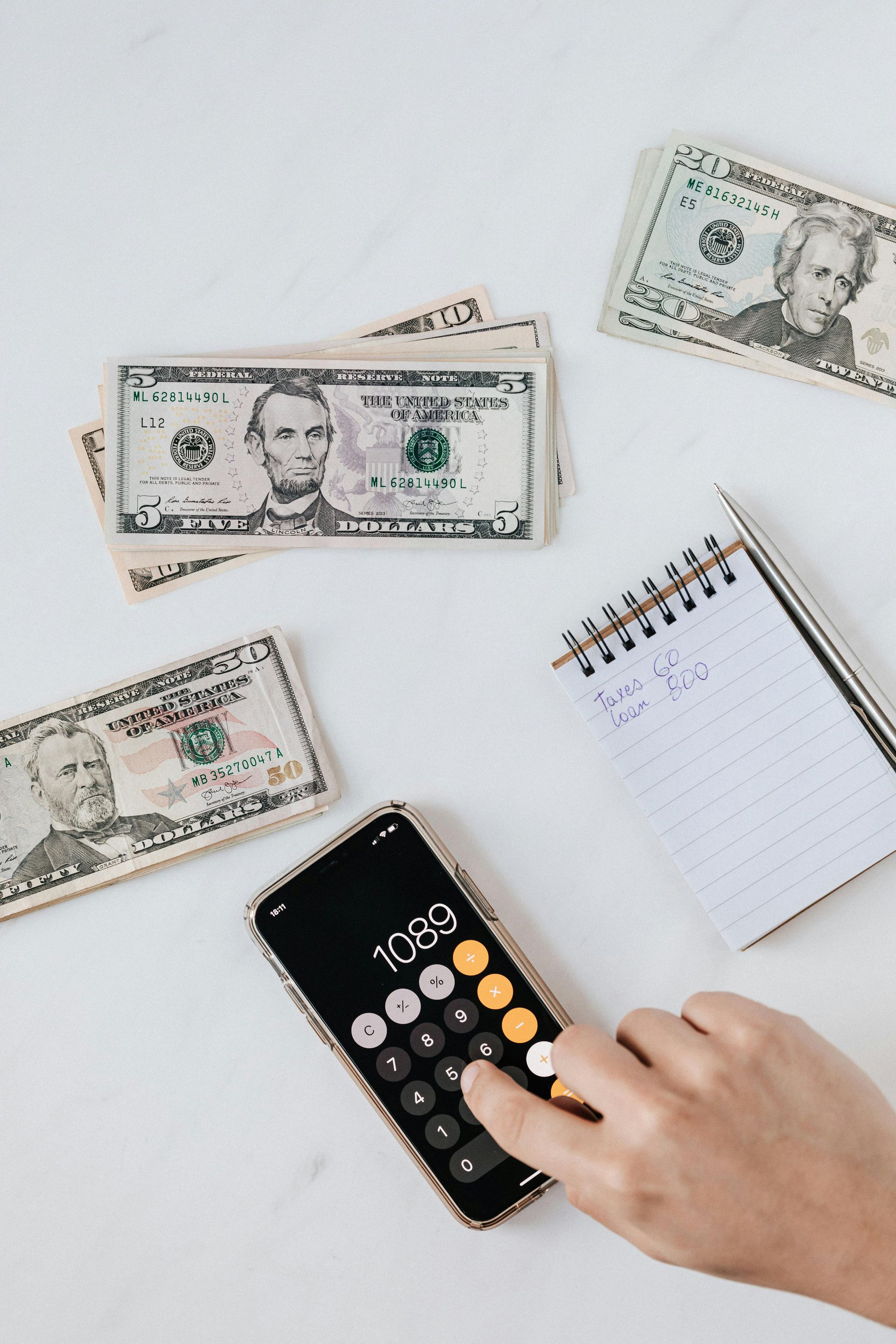 Person using a calculator surrounded by cash and a notepad on a white surface.