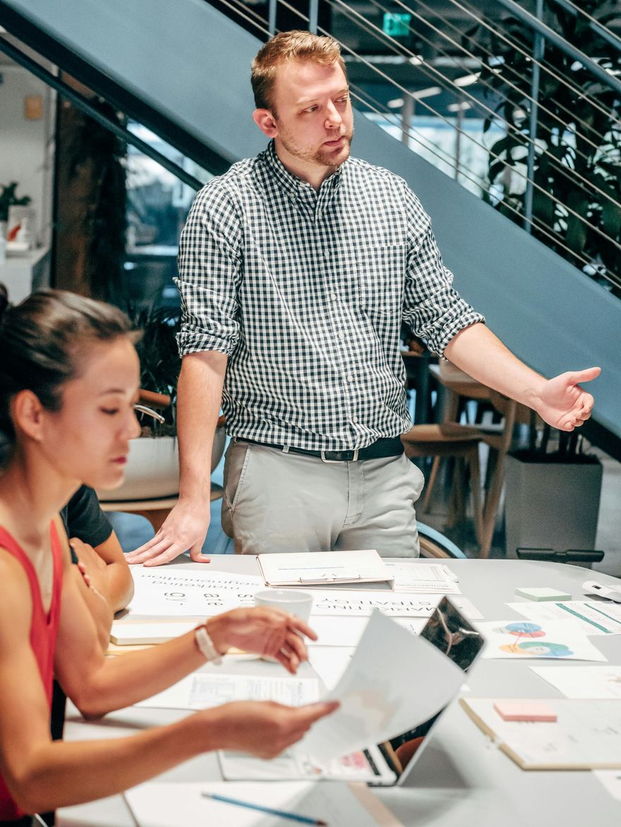 Man in checkered shirt gestures during a meeting; woman looks at documents, laptop open. Office setting.
