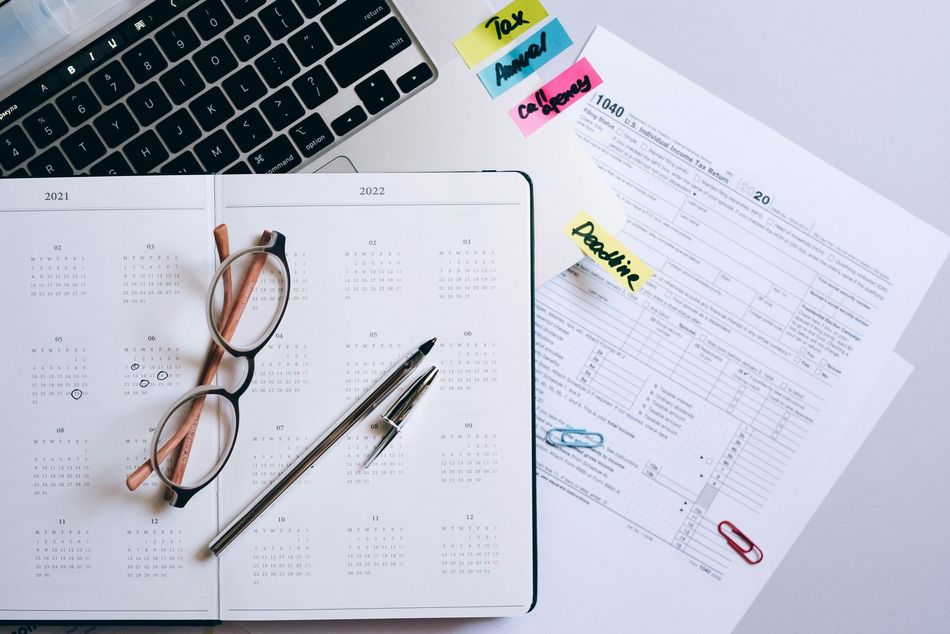 Laptop, planner, glasses, pen, and tax forms on a white surface with sticky notes.