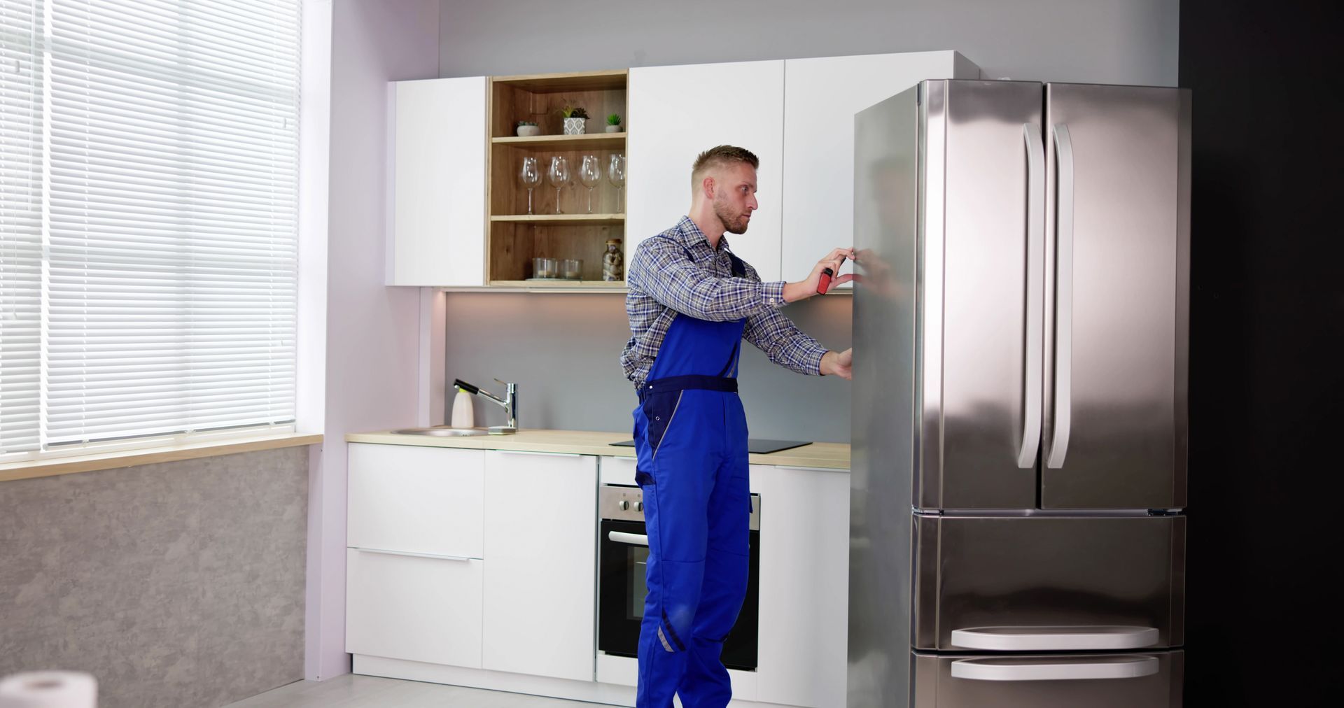 Technician in blue uniform performing Samsung fridge repair in modern white kitchen.