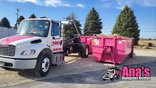 White tow truck loading a pink dumpster trailer in a sunny parking lot with trees and clear sky