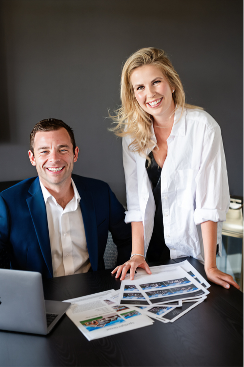 Two coworkers in business attire smiling at a desk with a laptop and printed design layouts.