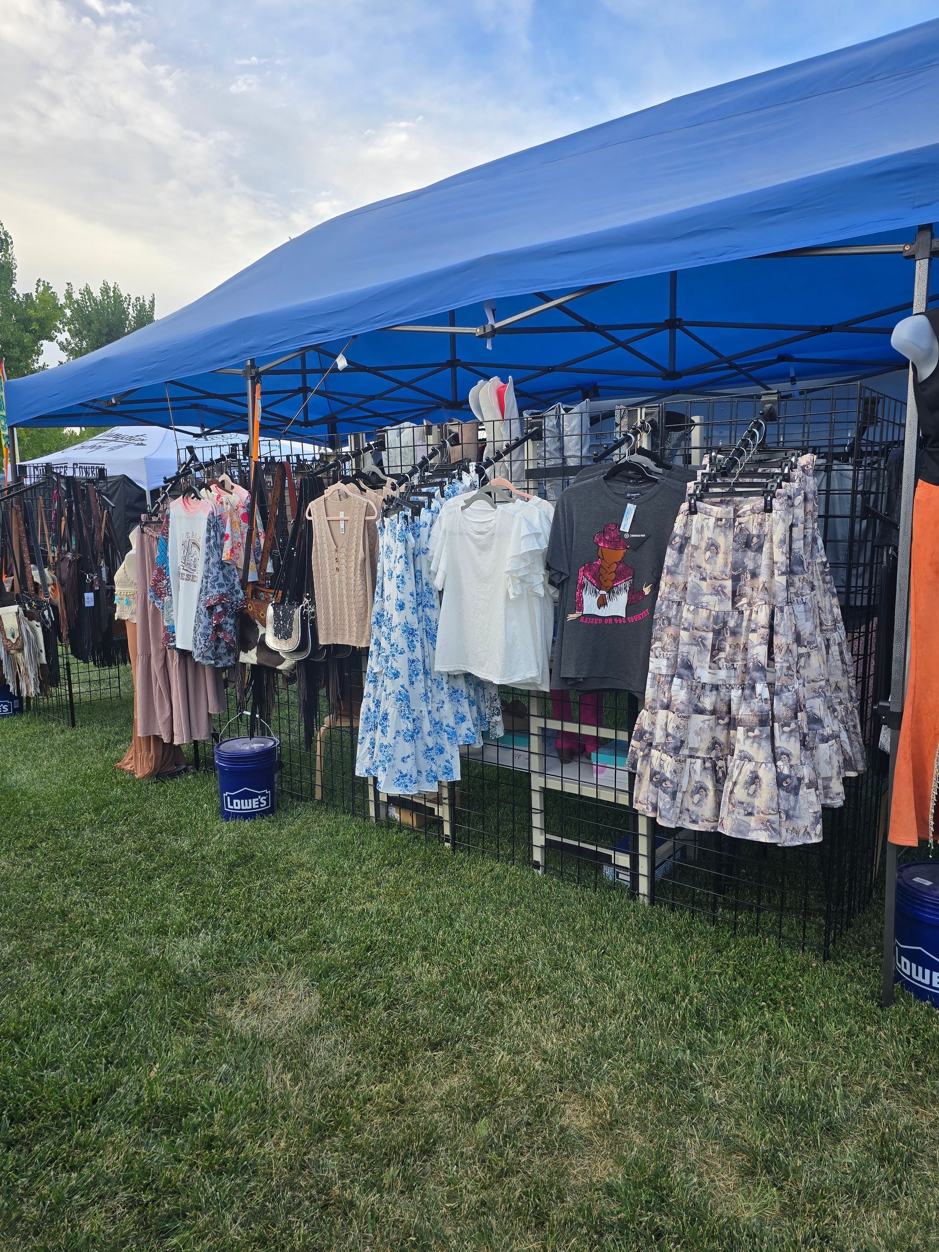 Clothing display under a blue tent at an outdoor market. Dresses, shirts, and skirts hang on racks.