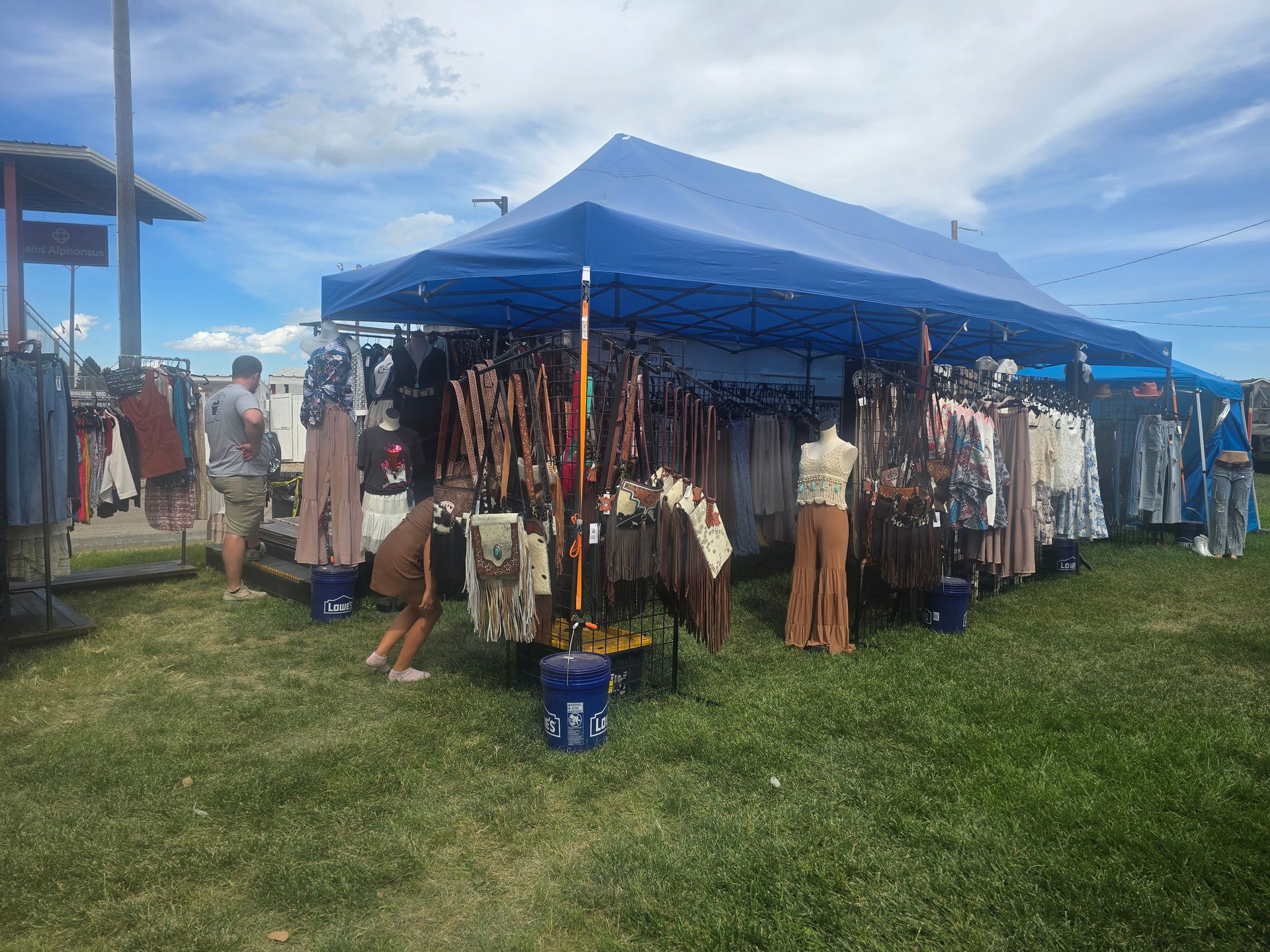 A clothing market stall under a blue canopy on a grassy field. People browse clothing on racks.
