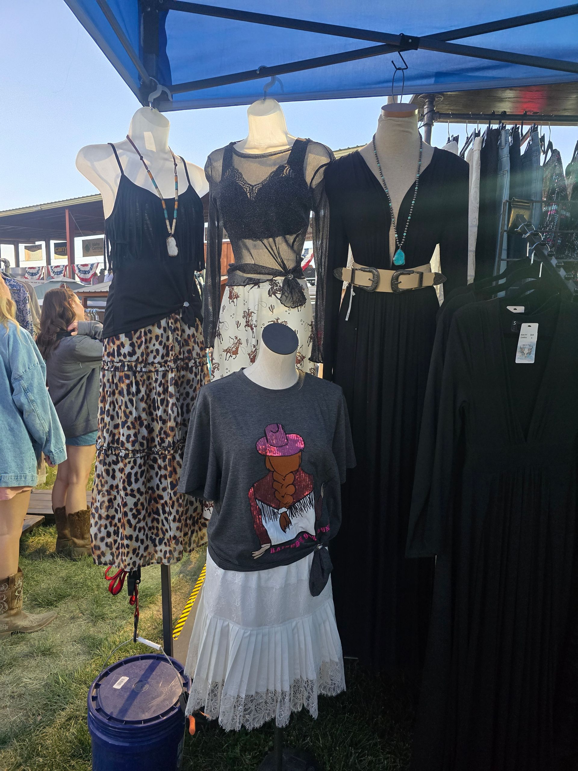 Clothing display on mannequins at outdoor market. Black, patterned, and white outfits hang.