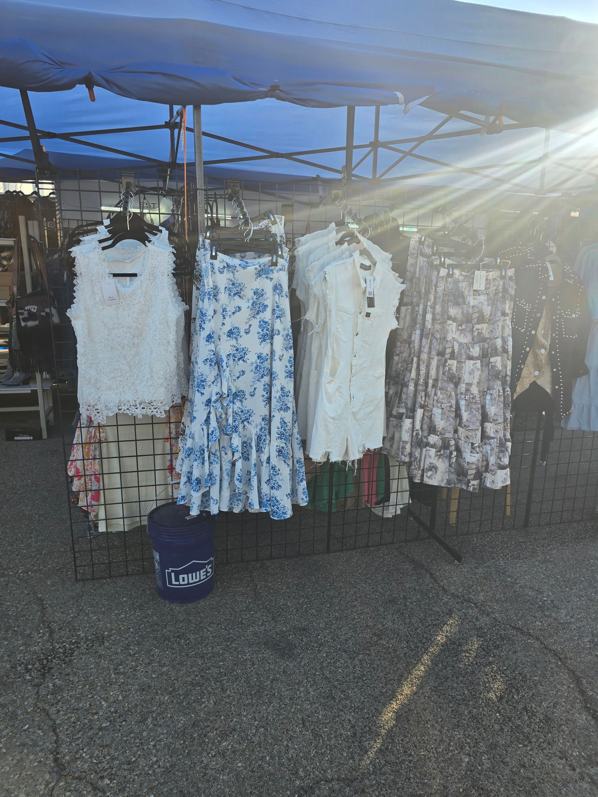 Clothing rack under a blue tarp; dresses in white, blue floral, and patterned fabrics. Outdoors, sunny.