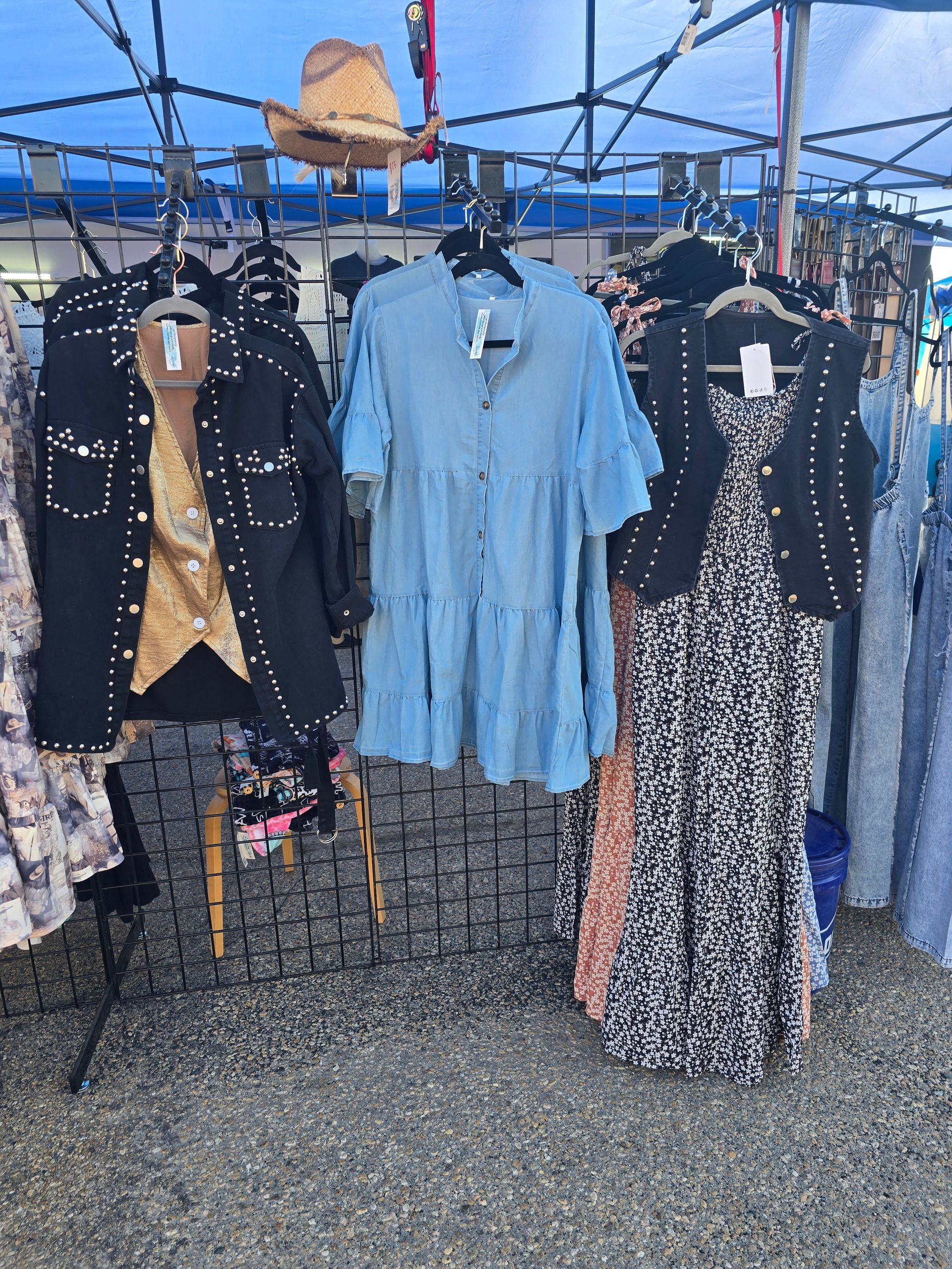 Clothing display at an outdoor market featuring jackets and dresses in blue, black, and patterns, under a blue canopy.