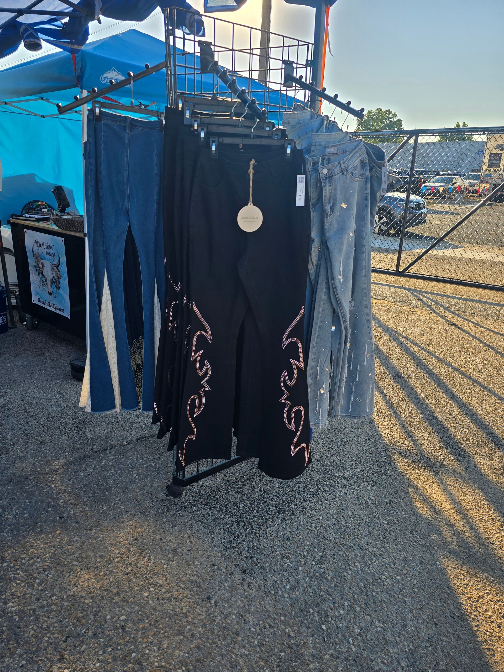 Jeans hanging on a rack at an outdoor market, black pants with pink designs, blue jeans, gravel ground.
