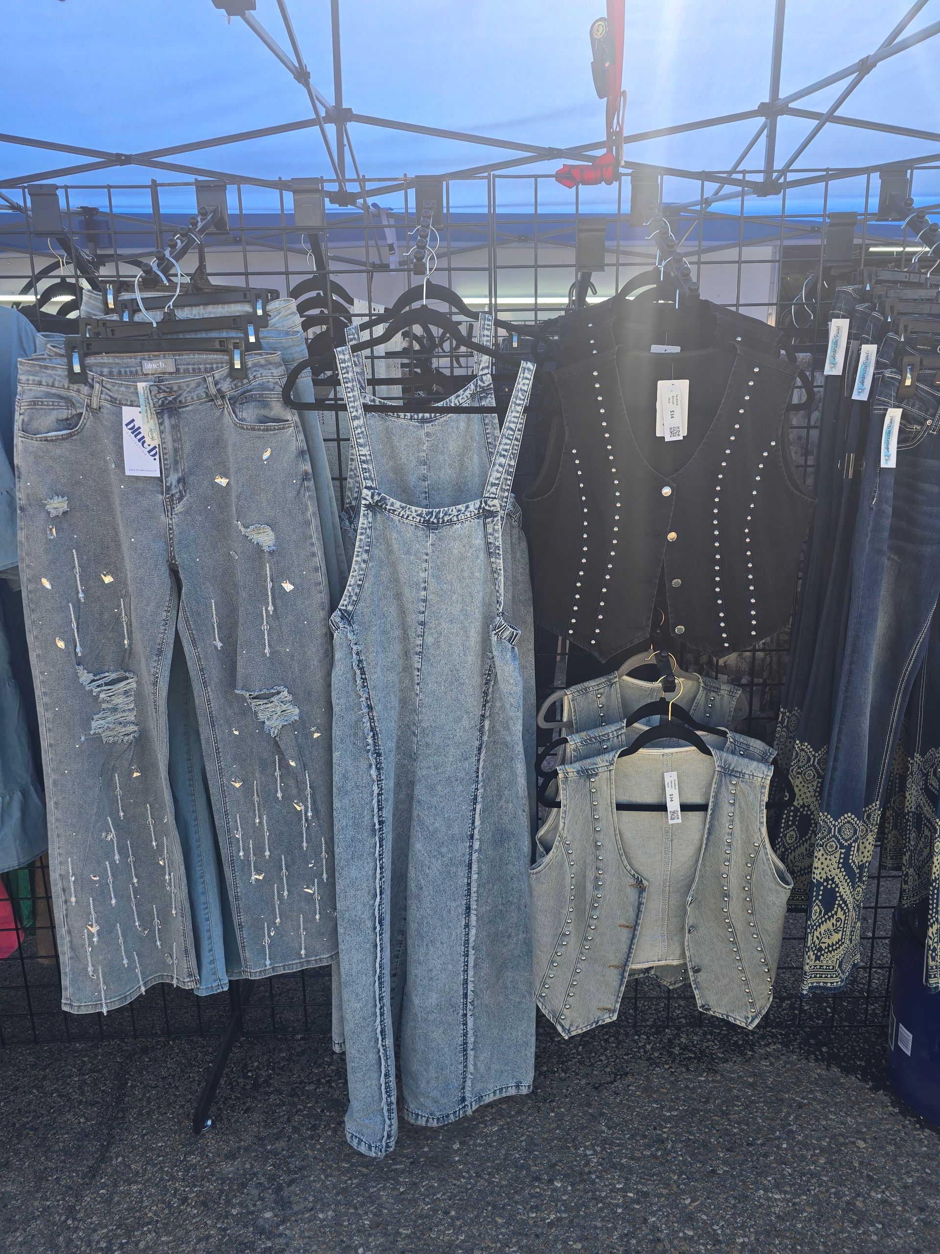 Denim clothing display: jeans, overalls, vest, and jacket with sparkle accents on hangers in a marketplace.