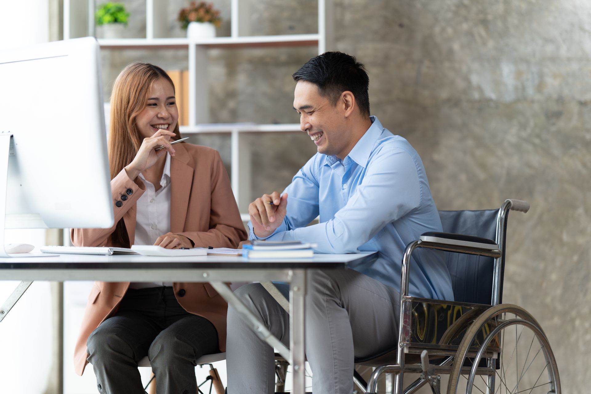 Two coworkers smiling at a table, one in a wheelchair, discussing paperwork in a bright office