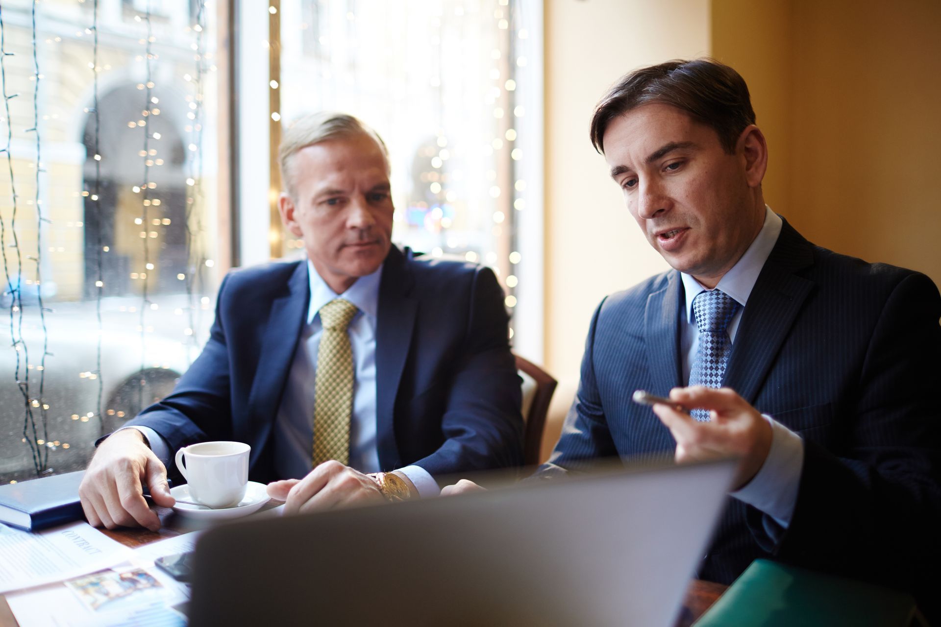 Two businesspeople in suits discussing at a table, with a laptop and coffee cups by a window.