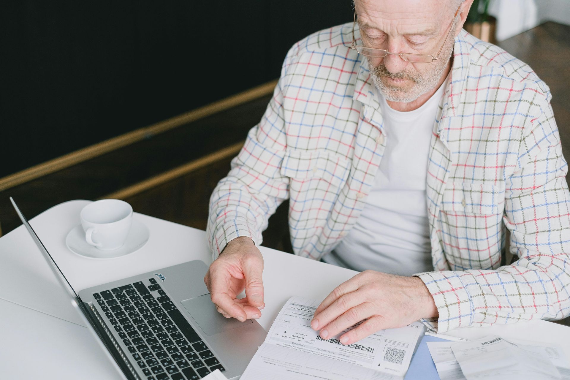 Person in a plaid shirt writing on papers at a table beside a laptop and coffee cup