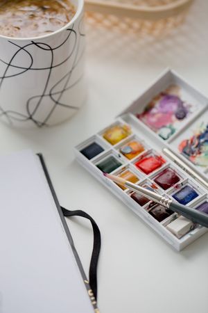 Un set di acquerelli accanto a un album da disegno e una tazza su un tavolo.